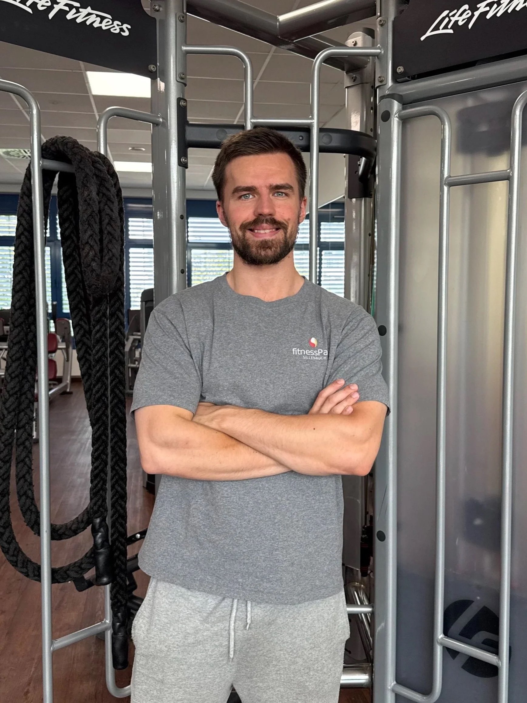 Junge Mann, stehend in einem Fitnessstudio, lächelnd, mit verschränkten Armen, trägt ein graues T-Shirt mit einem Fitnessstudio-Logo, im Hintergrund Fitnessgeräte und schwarze Bänder.