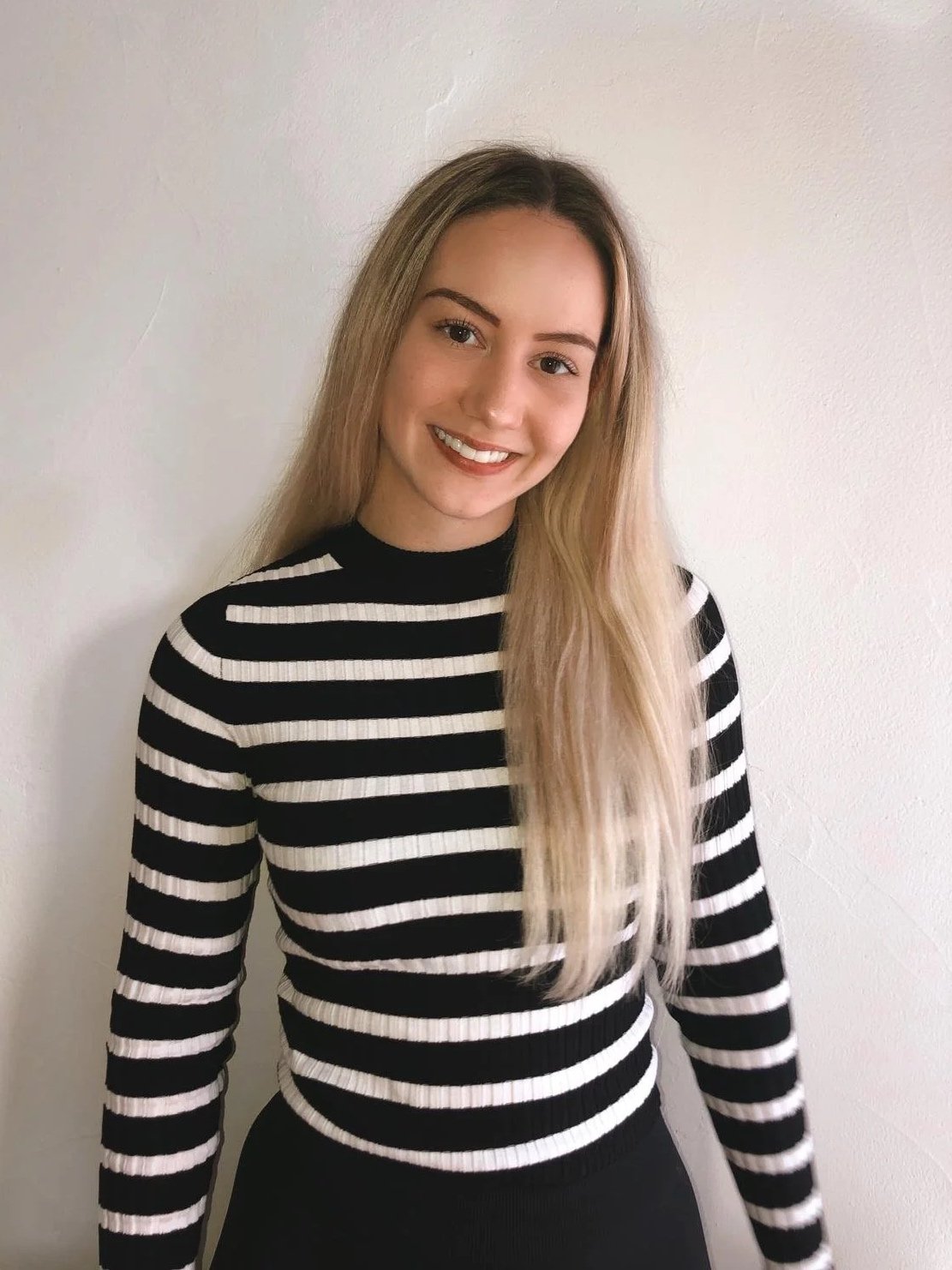 Junge Frau mit langem, blondem Haar, lächelnd, trägt ein schwarzes und weiß gestreiftes Oberteil, steht vor weißer Wand.