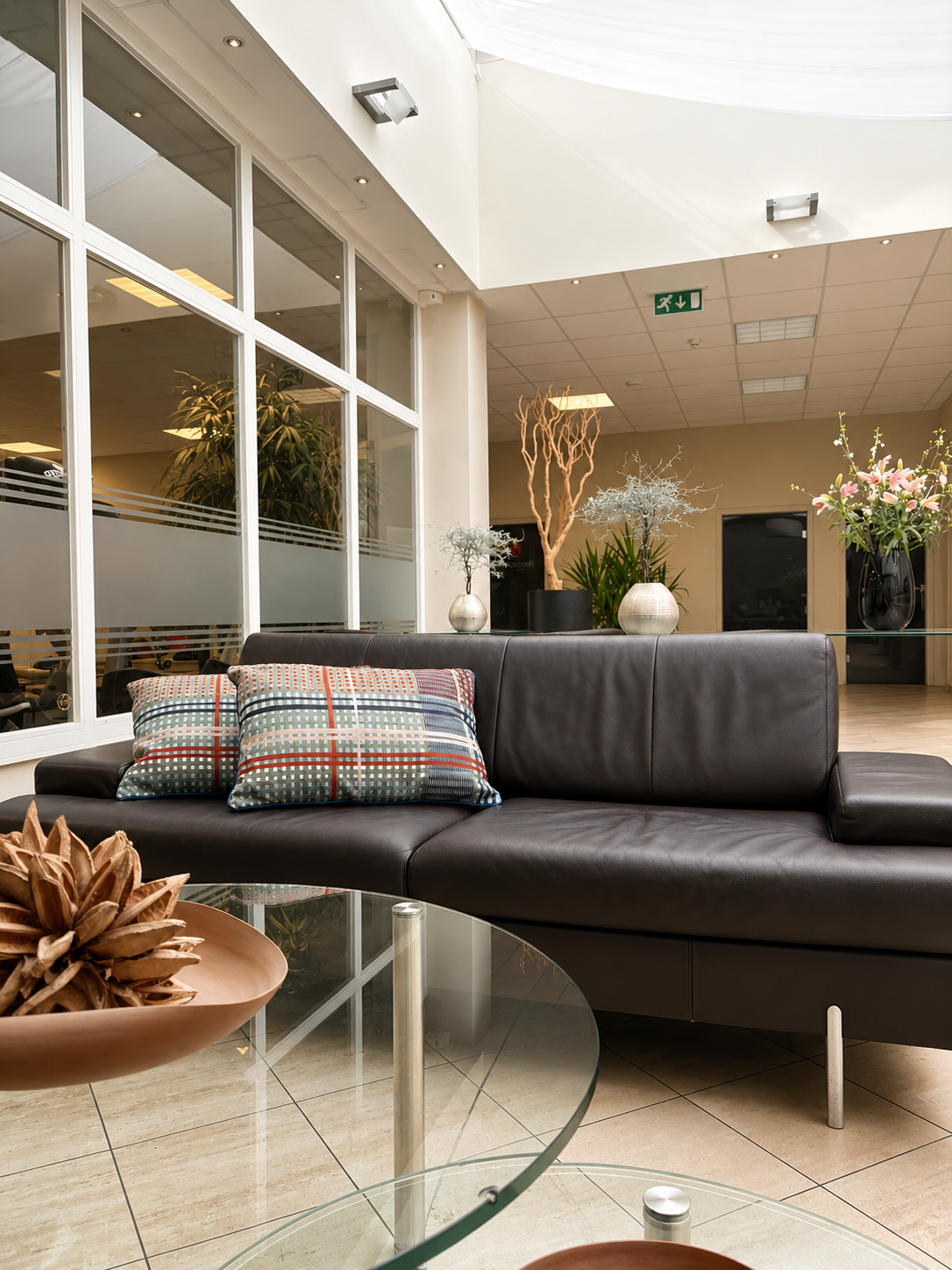 Ein modernes Wartezimmer mit schwarzem Ledersofa, bunten Kissen, Glas-Tisch, Pflanzen und Blumen an der Wand, helle Raumgestaltung.