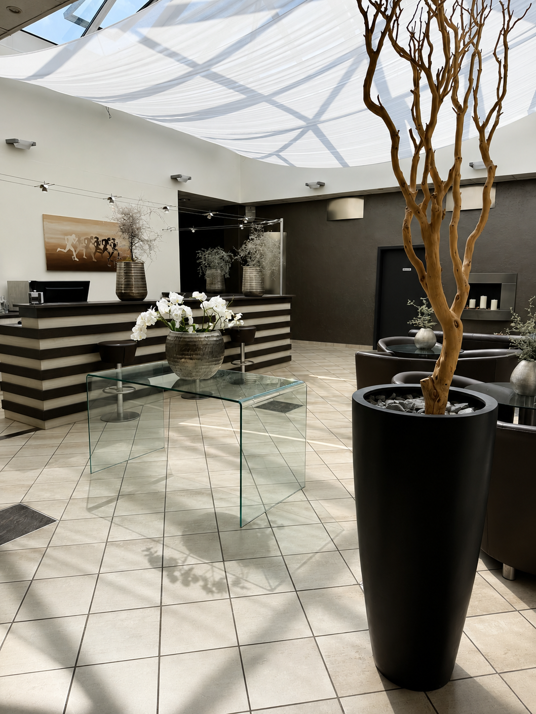 Innenaufnahme eines stilvoll eingerichteten Wartezimmers mit großen schwarzen Pflanzgefäßen, weißen Orchideen, einem Glastisch, einer Bar sowie weiteren Pflanzen und Dekorationen.