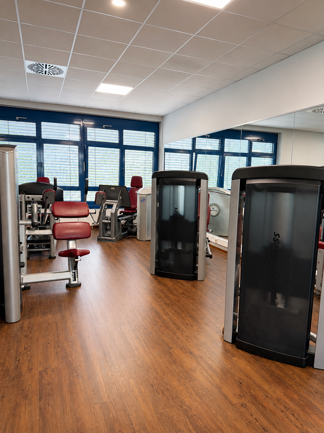 Fitnessstudio mit verschiedene Trainingsgeräte, Holzfußboden, große Fenster mit blauen Rahmen und Rollladen, spiegelnde Wand auf der rechten Seite.