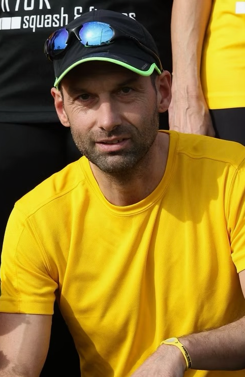 Ein Mann in gelbem T-Shirt und schwarzer Kappe mit Sonnenbrille sitzt, im Hintergrund sind andere Personen, vermutlich bei einer Sportveranstaltung.