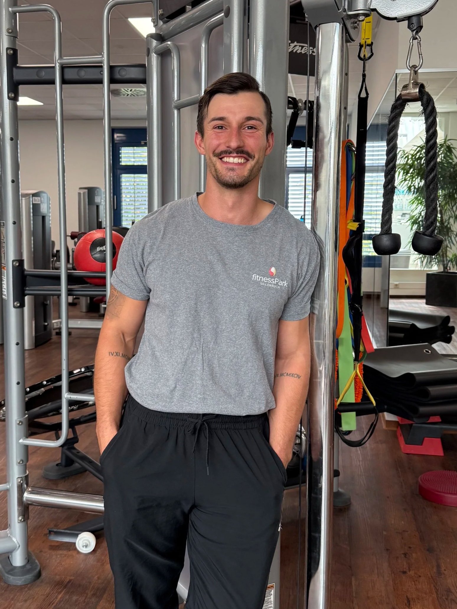 Ein Mann in Sportkleidung steht lächelnd in einem Fitnessstudio, gegenüber einem Kraftgerät.