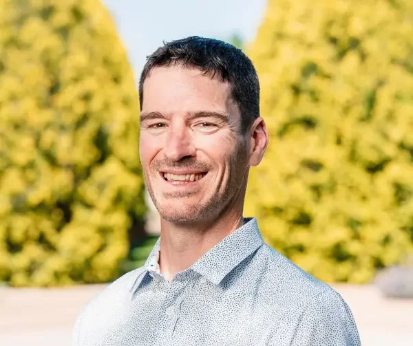 A smiling man with short dark hair and a light patterned dress shirt, outdoor background with yellow trees.