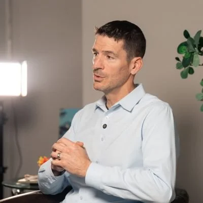 A man with short dark hair, wearing a light blue button-up shirt, stands with his hands clasped in front of him in a room with a plant and a light on the wall.