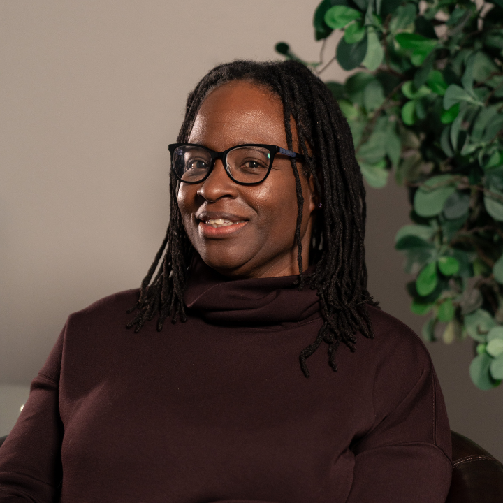 A woman with long dreadlocks and glasses smiling while seated indoors with a potted plant in the background.