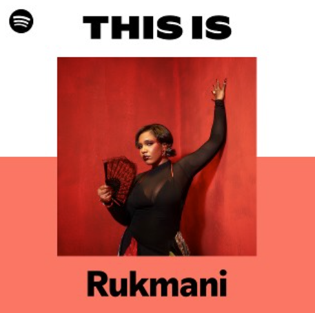 A woman dressed in black holding a fan, standing in front of a red wall with a white and red background. Text above her reads 'THIS IS' and below reads 'Rukmani' in bold black letters.