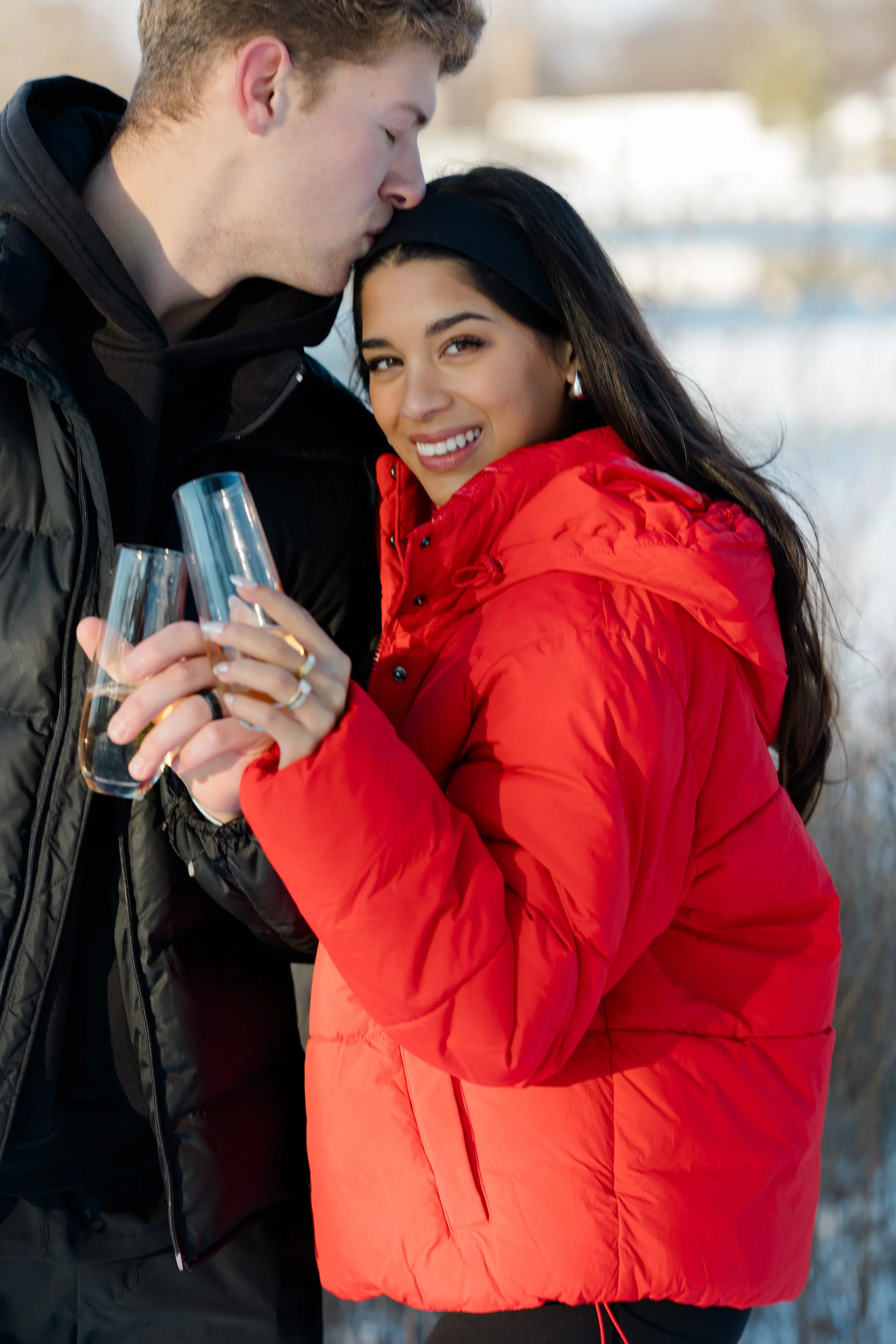 A young man kissing a young woman on the head outdoors in winter while holding two champagne glasses. She is smiling, wearing a red jacket and black headband, and she has dark hair and earrings.
