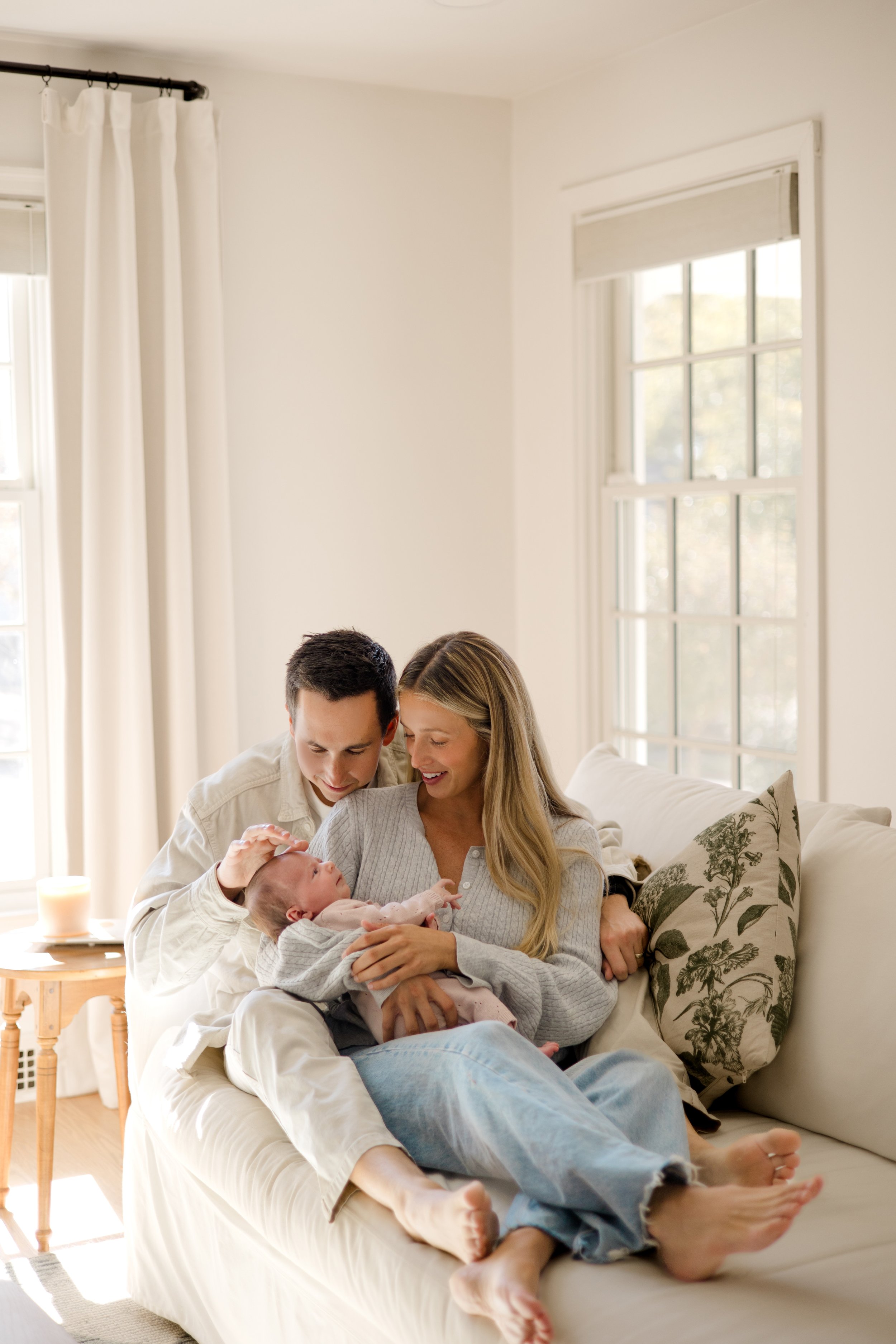 Family in a cozy living room with white walls and large windows, sitting on a sofa, holding a newborn baby, with a woman and man smiling and looking at the baby.