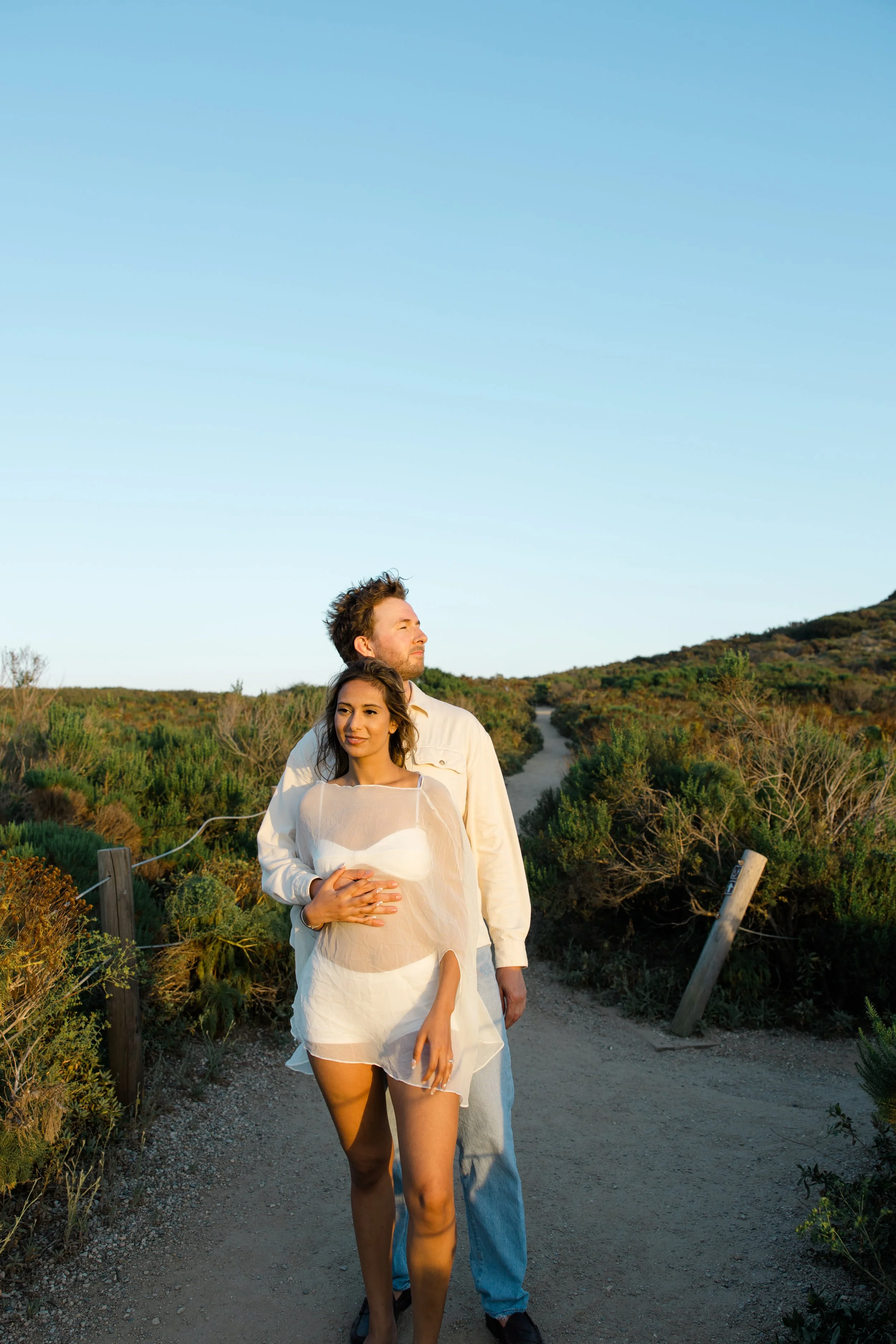 A pregnant woman in a semi-transparent white dress holding her belly, walking on a dirt path with a man behind her who is gently holding her. They are in a scenic outdoor setting with greenery and a hill in the background under clear blue sky at suns