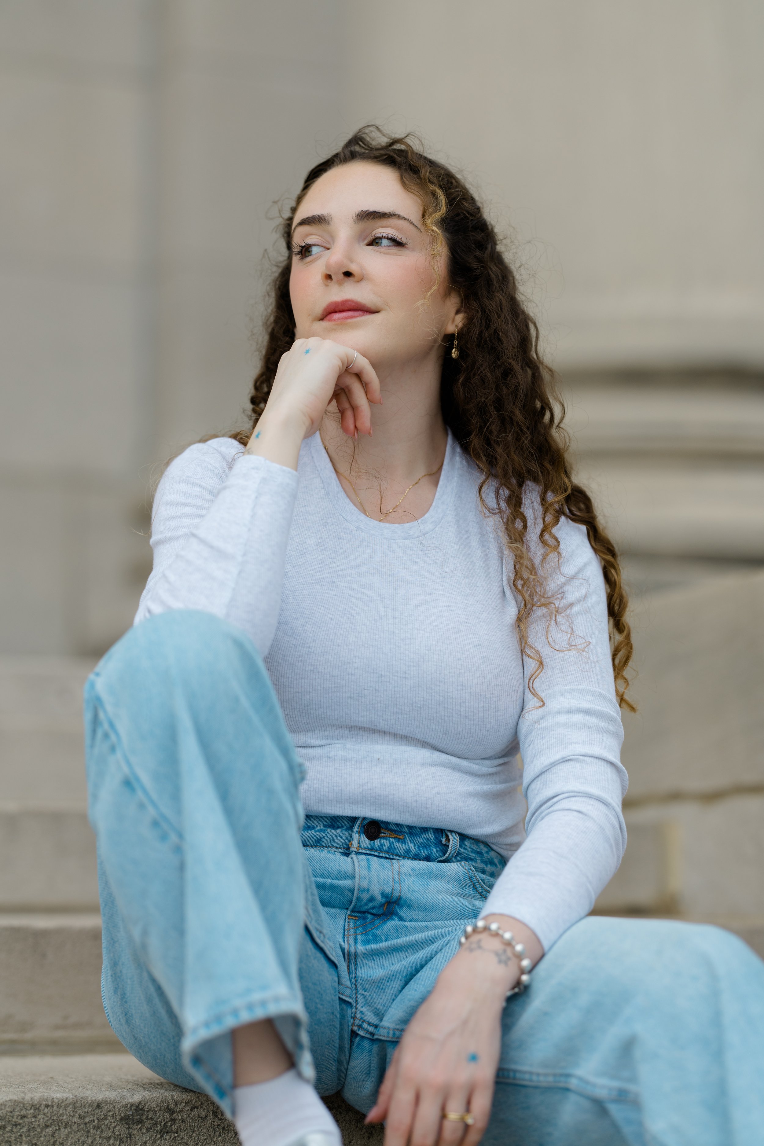 A young woman with long, curly brown hair, wearing a light gray long-sleeve shirt and light blue jeans, sitting on steps outside with her chin resting on her hand, looking to the side with a thoughtful expression.