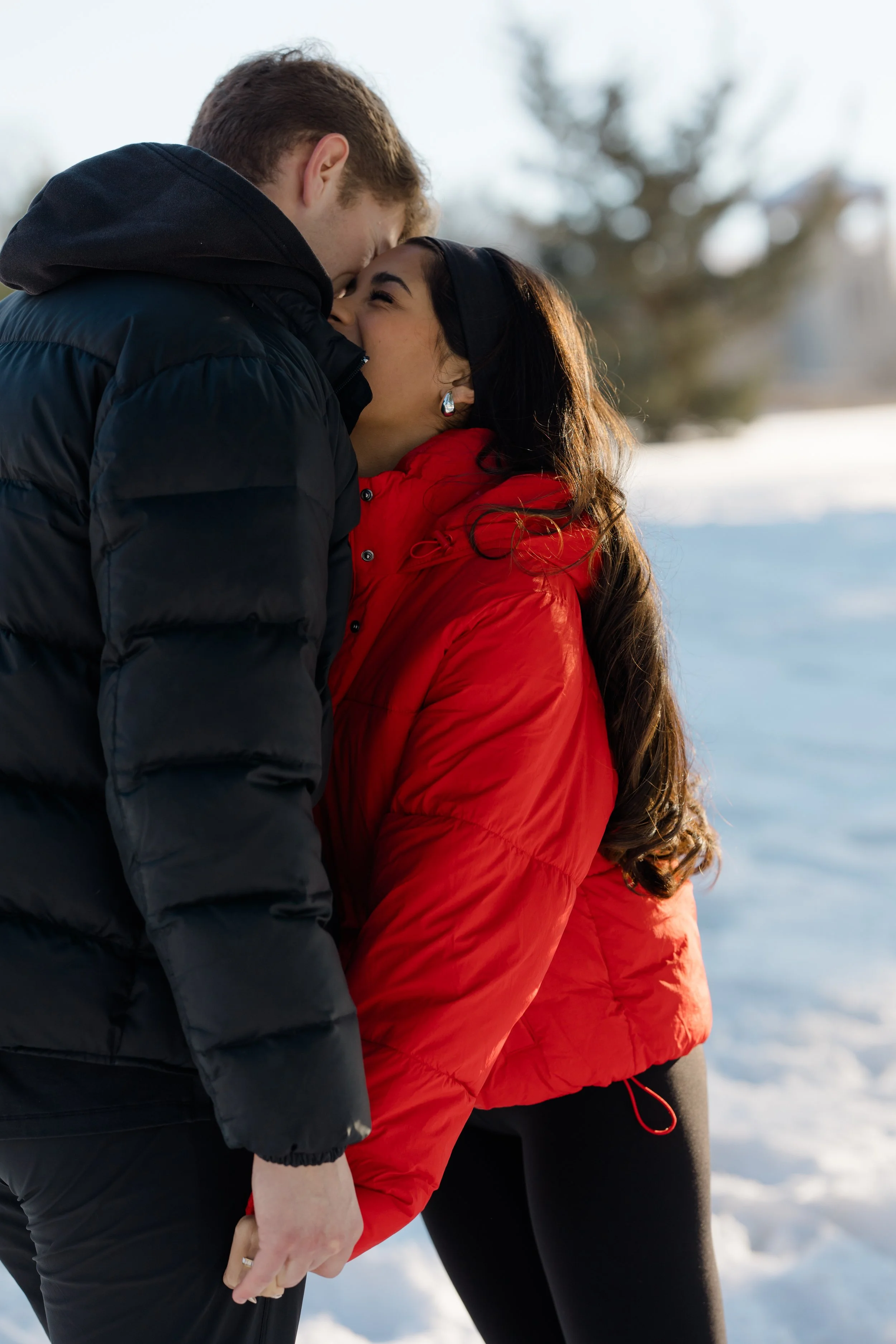 A couple in winter clothing holding hands outdoors in a snowy landscape, with their foreheads touching and smiling.