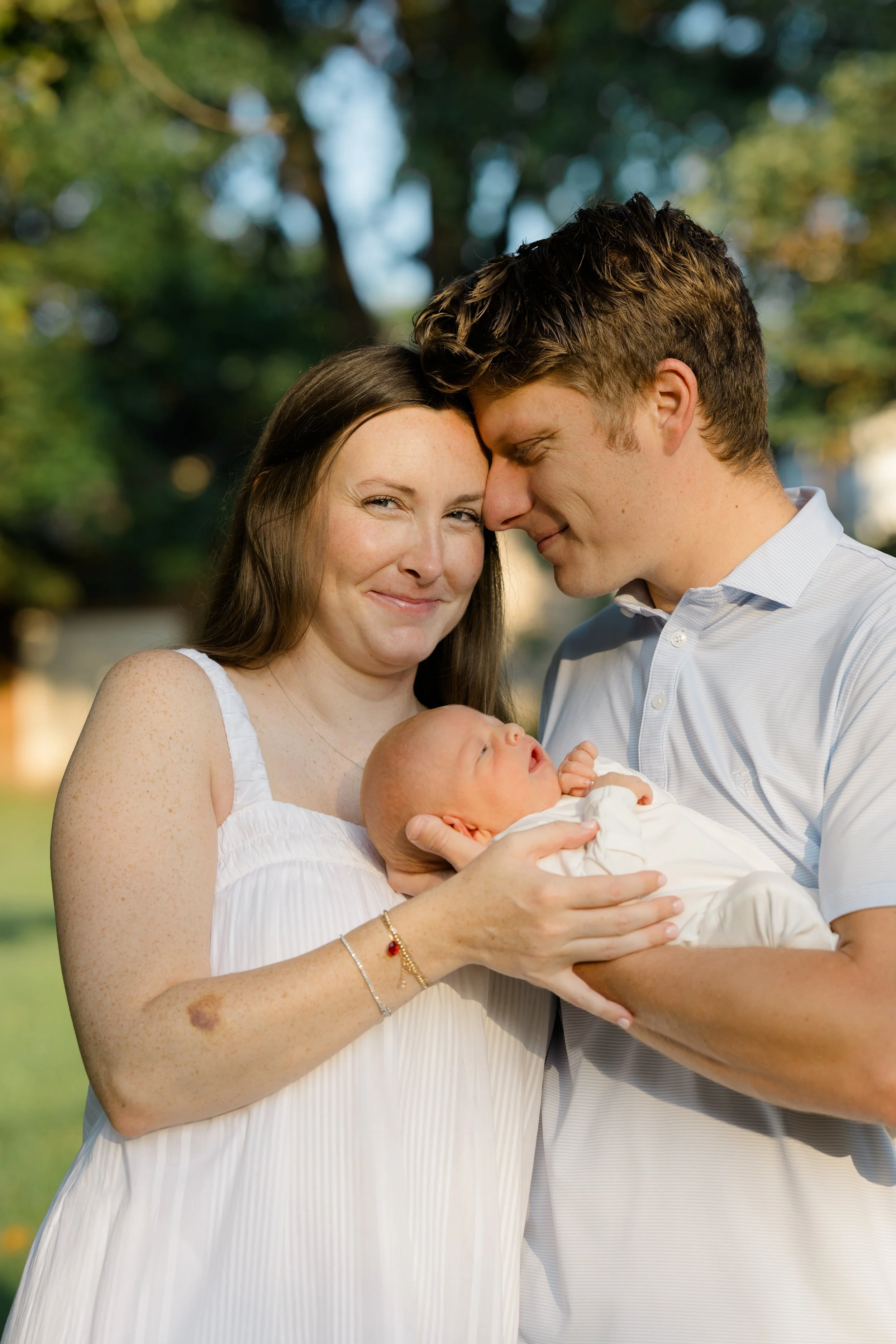 A happy couple holding a newborn baby outdoors during daytime. The woman has long brown hair, wears a white dress, and smiles affectionately, while the man with curly brown hair and a light blue shirt gently touches the baby's head, who is dressed in