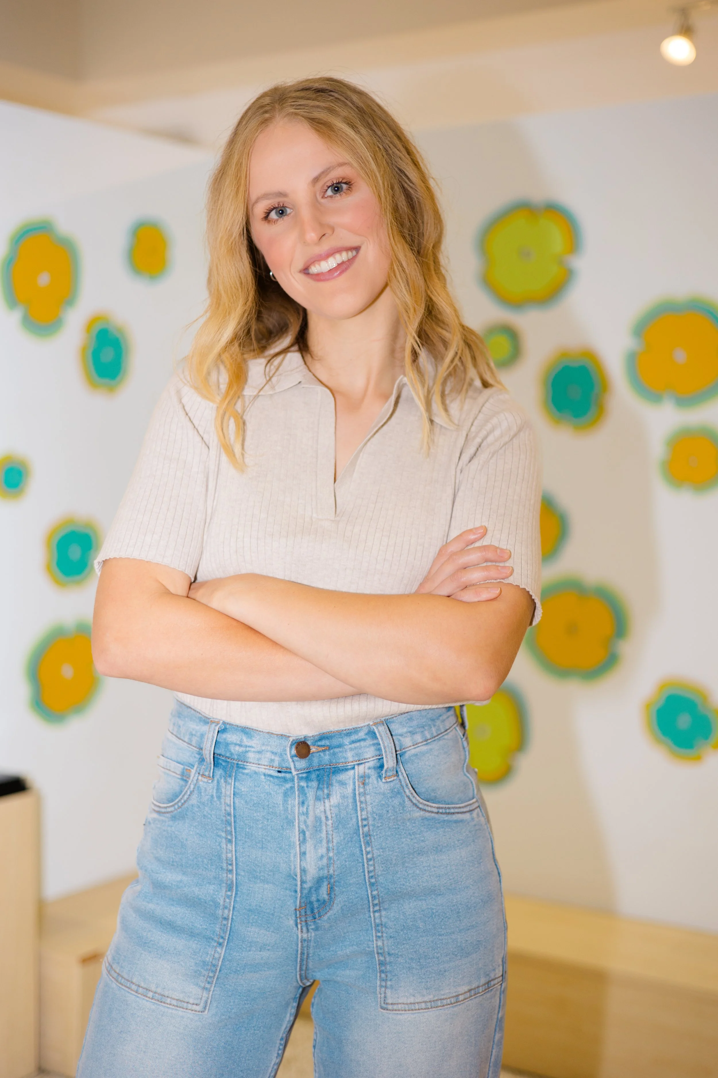 A woman with blonde hair smiling and standing with arms crossed in a bright room with colorful flower decorations on the wall.