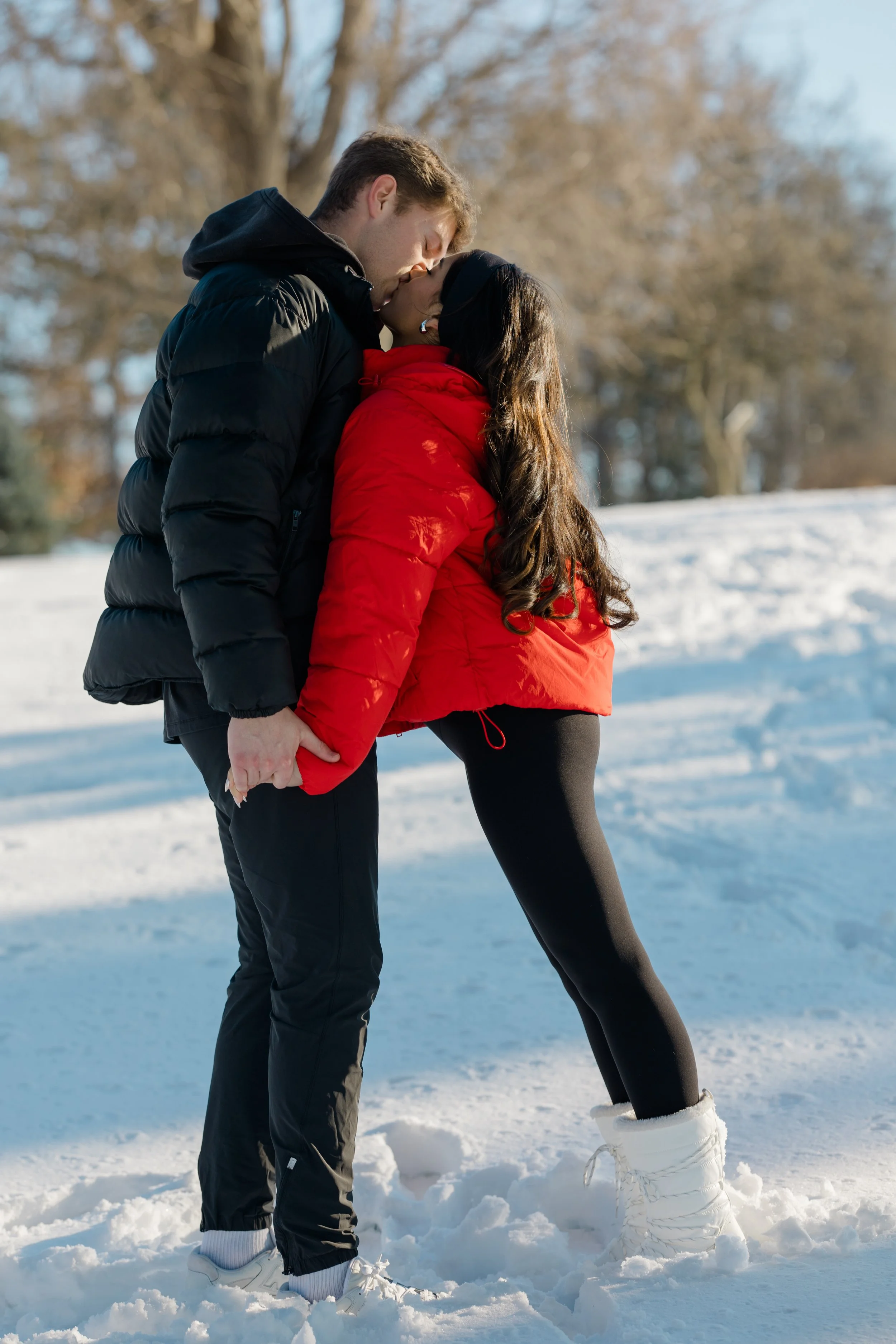A couple in winter clothing sharing a kiss outdoors in a snowy landscape with trees in the background.