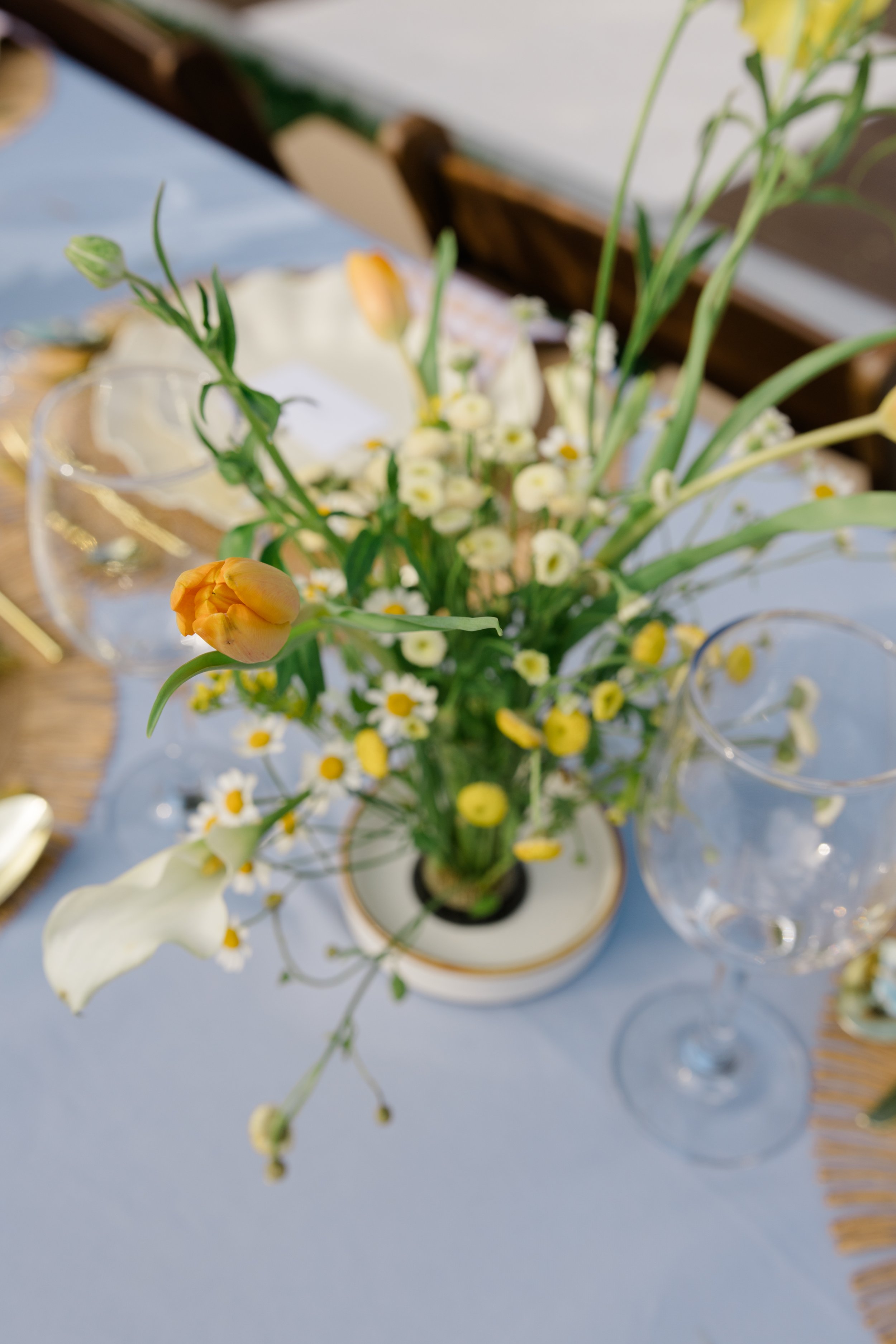 A floral centerpiece with white and yellow flowers in a vase on a table, set for a meal with glasses and a plate, in a well-lit room.