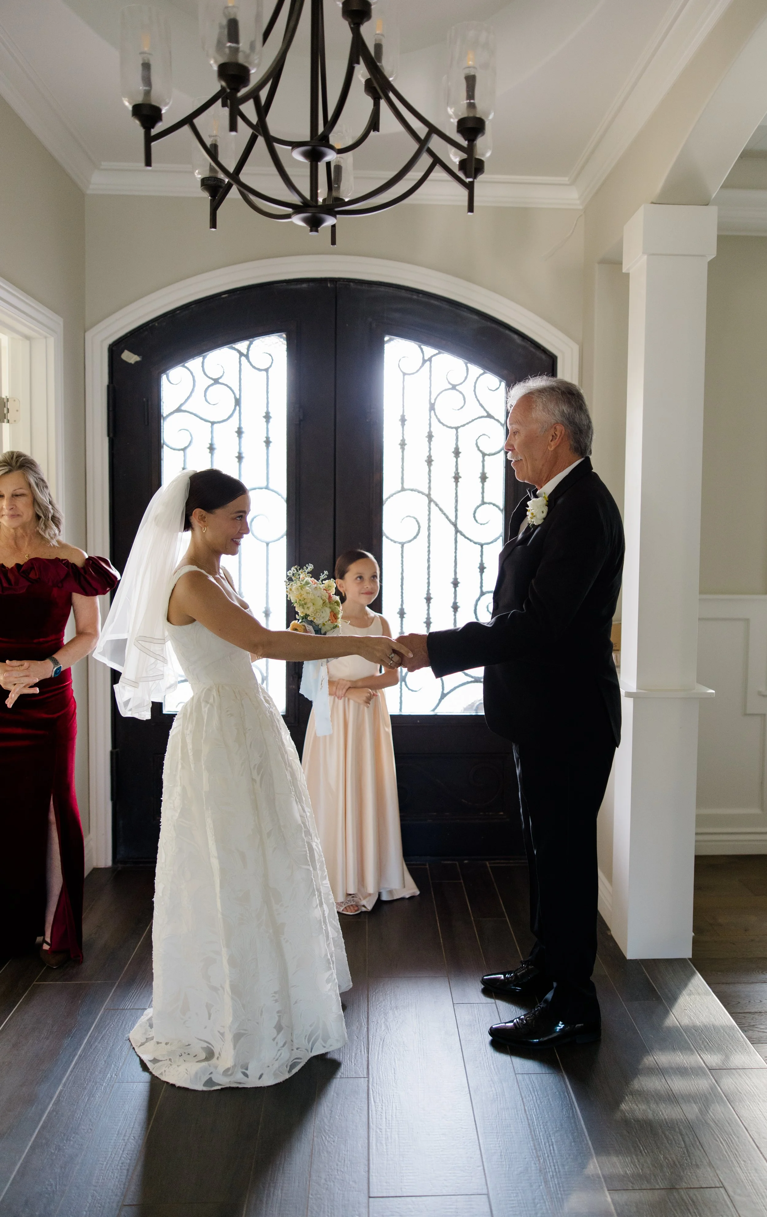 A bride and an older man, likely her father, holding hands during a wedding ceremony in a bright room with a large front door with decorative glass and black hardware. Two women stand in the background, one wearing a deep red gown and the other in a 