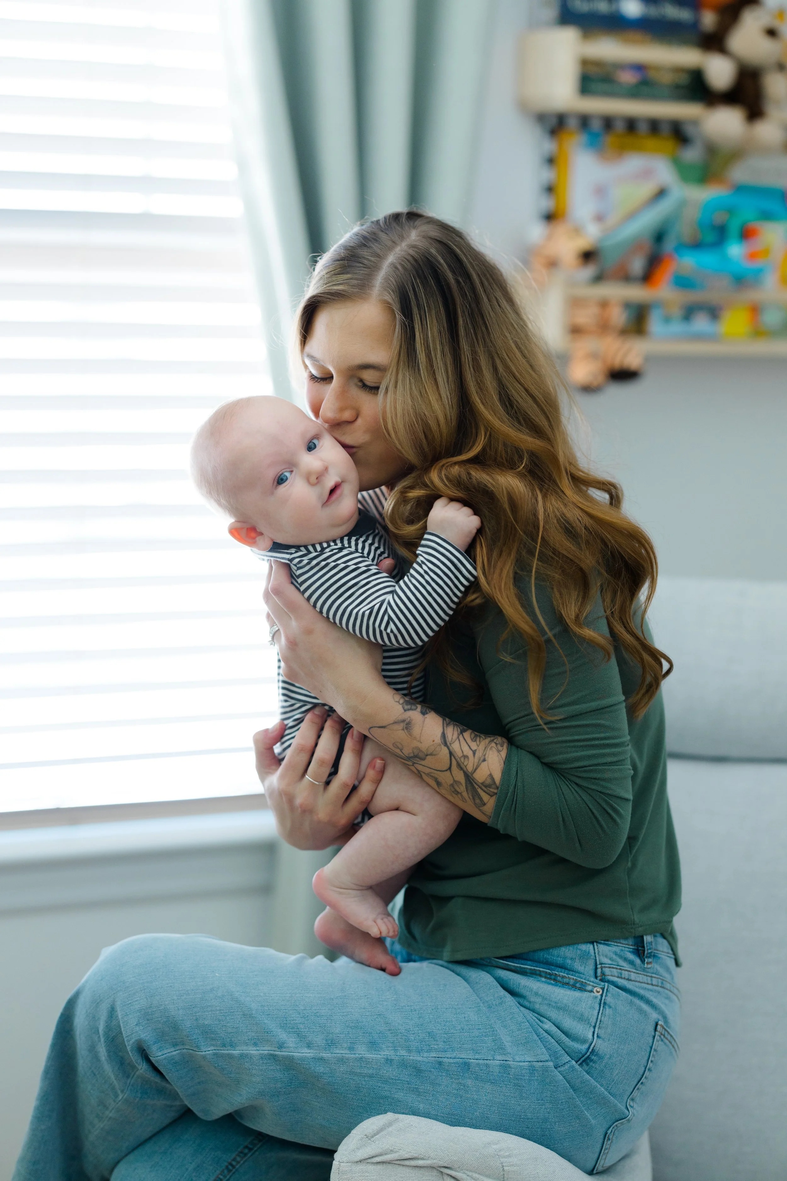 A woman with long, wavy red hair holding a baby with blue eyes, wearing a striped onesie, in a room with a window, curtains, and shelves with toys in the background.