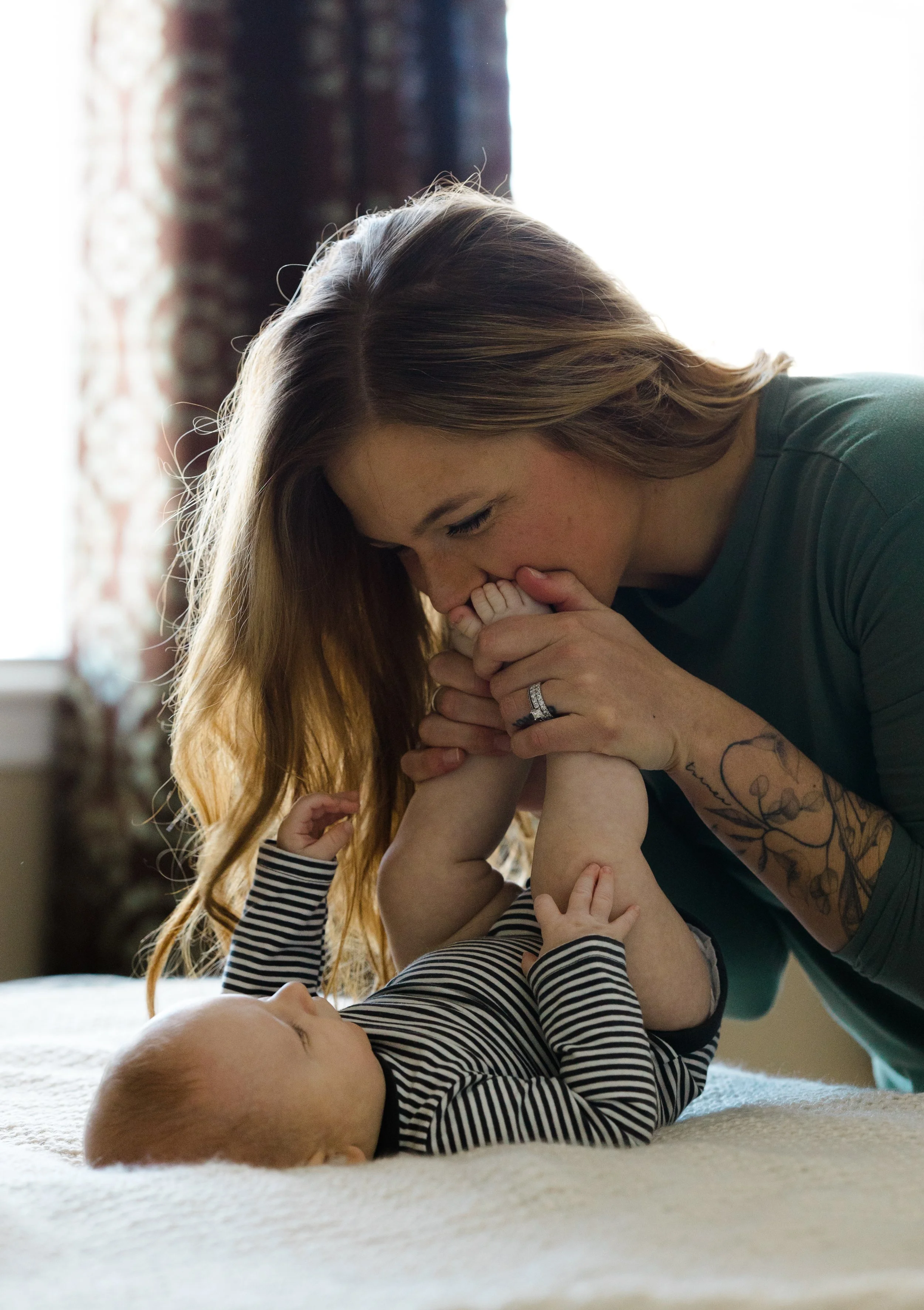 A woman with long, wavy hair playing with a baby who is lying on a bed. The woman is holding the baby's hands and bringing her close for a kiss. The baby has light skin, wears a striped shirt, and is reaching towards the woman. The background include
