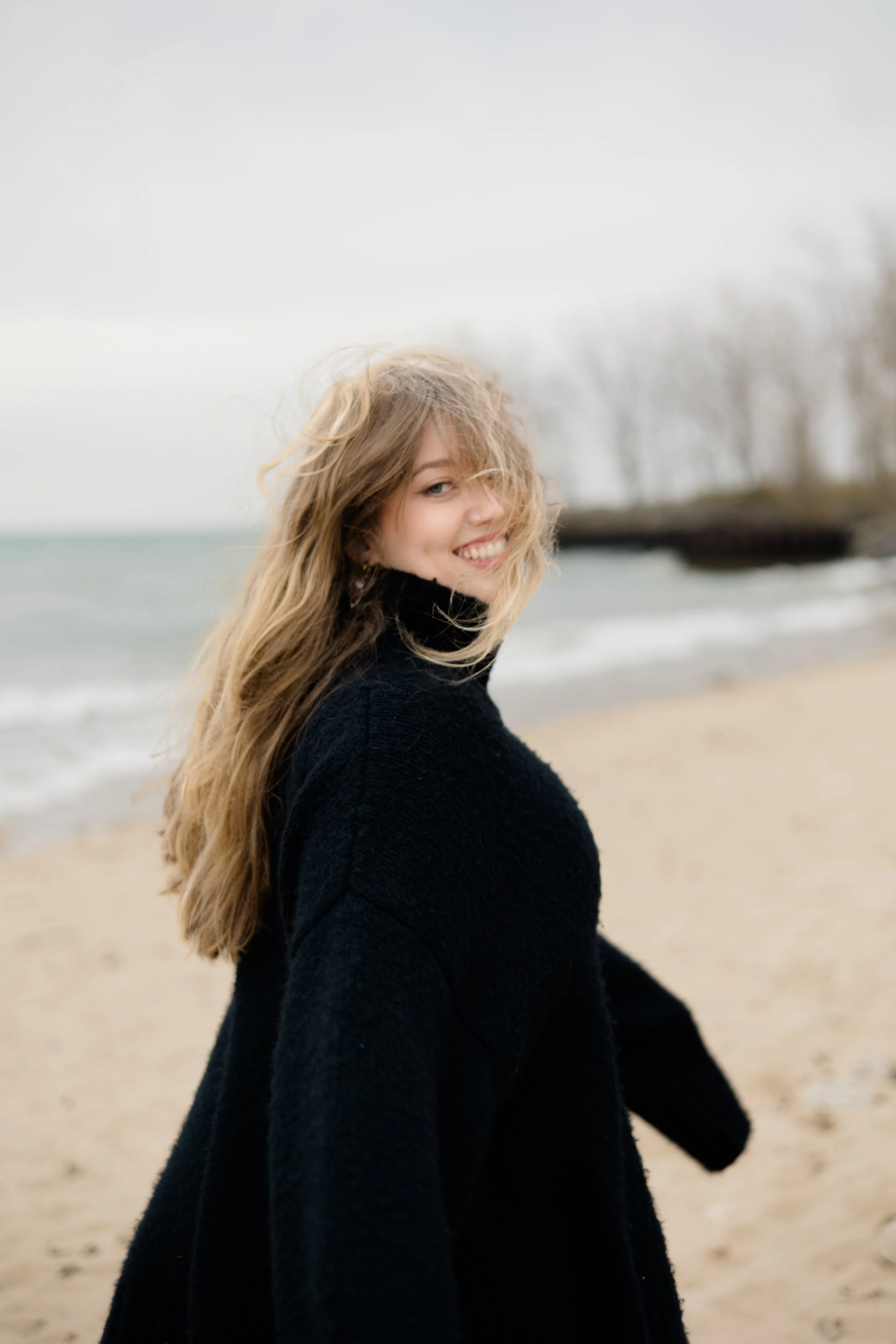 A woman with long wavy blonde hair standing on a beach, smiling and looking over her shoulder, with trees and a cloudy sky in the background.