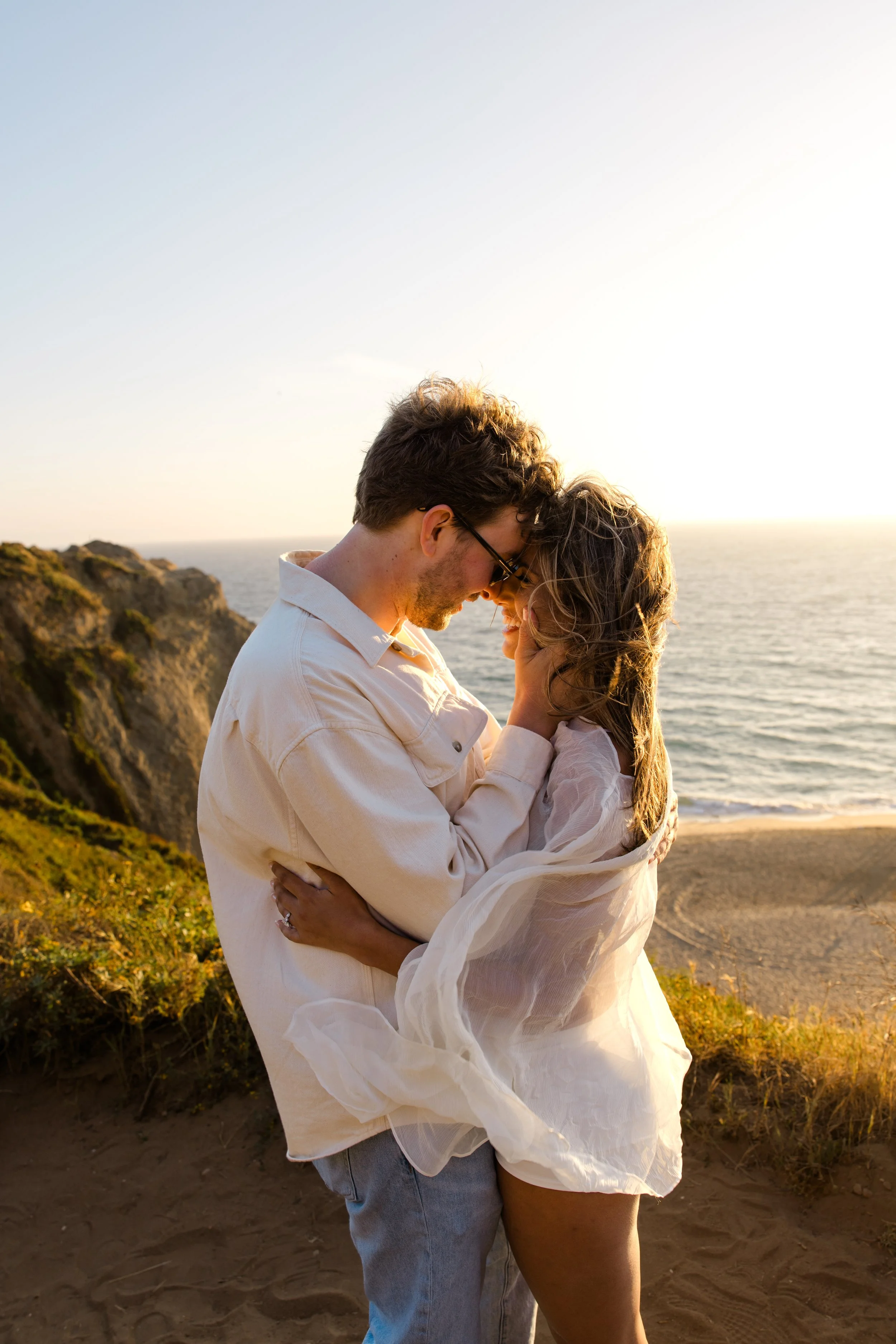 A couple embracing and touching foreheads on a beach at sunset, overlooking the ocean and cliffs.