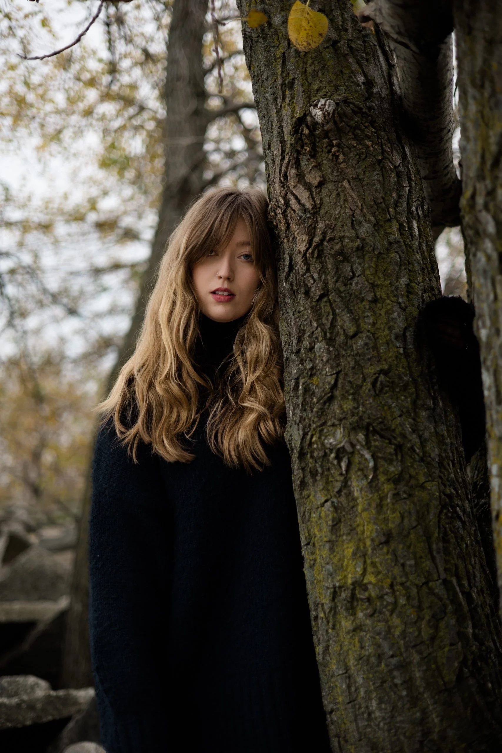 A young woman with long wavy hair and a serious expression, partially hidden behind a large tree in an outdoor setting with autumn foliage.