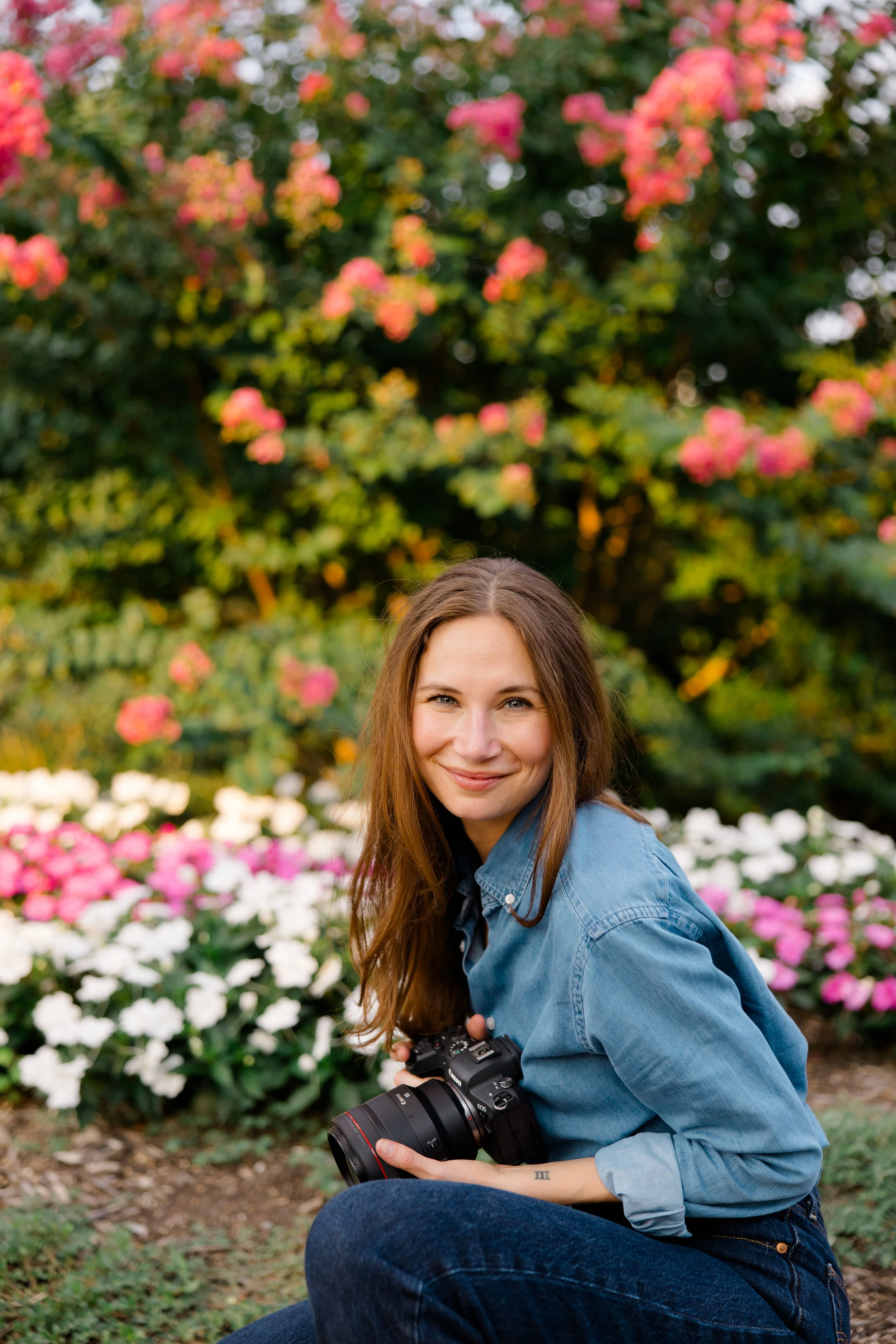 A young woman with long brown hair sitting outdoors, holding a camera, smiling, with colorful flowers and green foliage behind her.