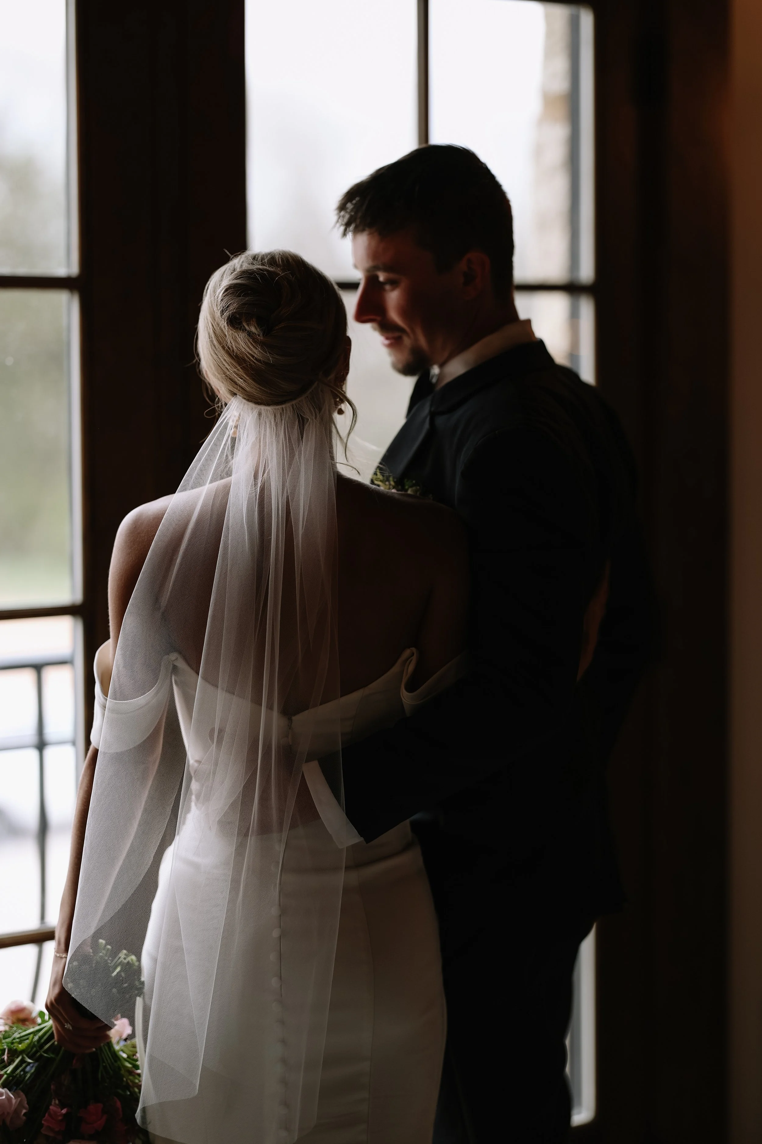 A bride and groom standing close to each other near a window, smiling and gazing at each other during their wedding.