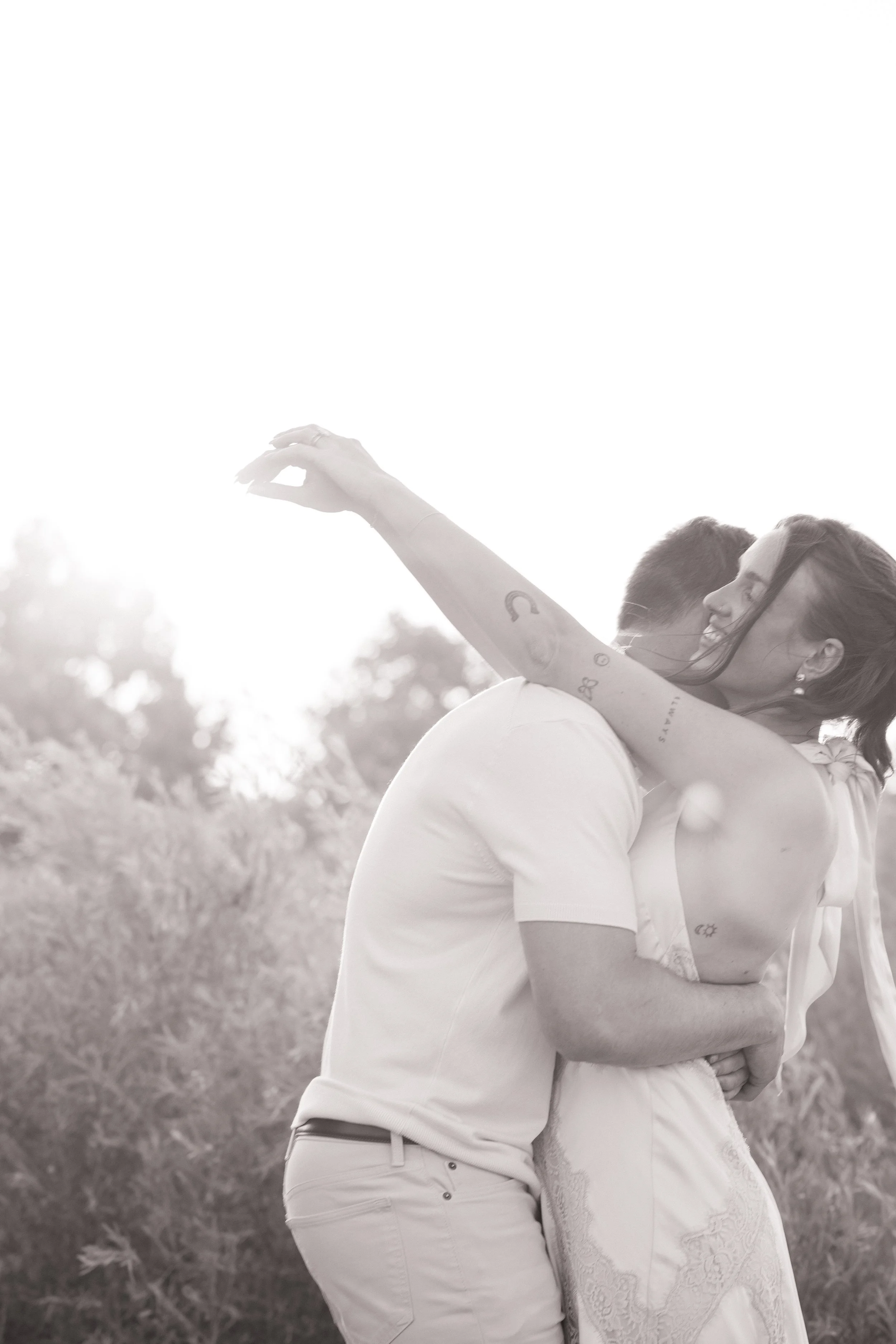 A couple is hugging outdoors during sunset, with the man’s face close to the woman's face. The woman is smiling with her eyes closed, and the man’s face is turned away. The woman has tattoos on her arm, and they are both dressed in light-colored clot