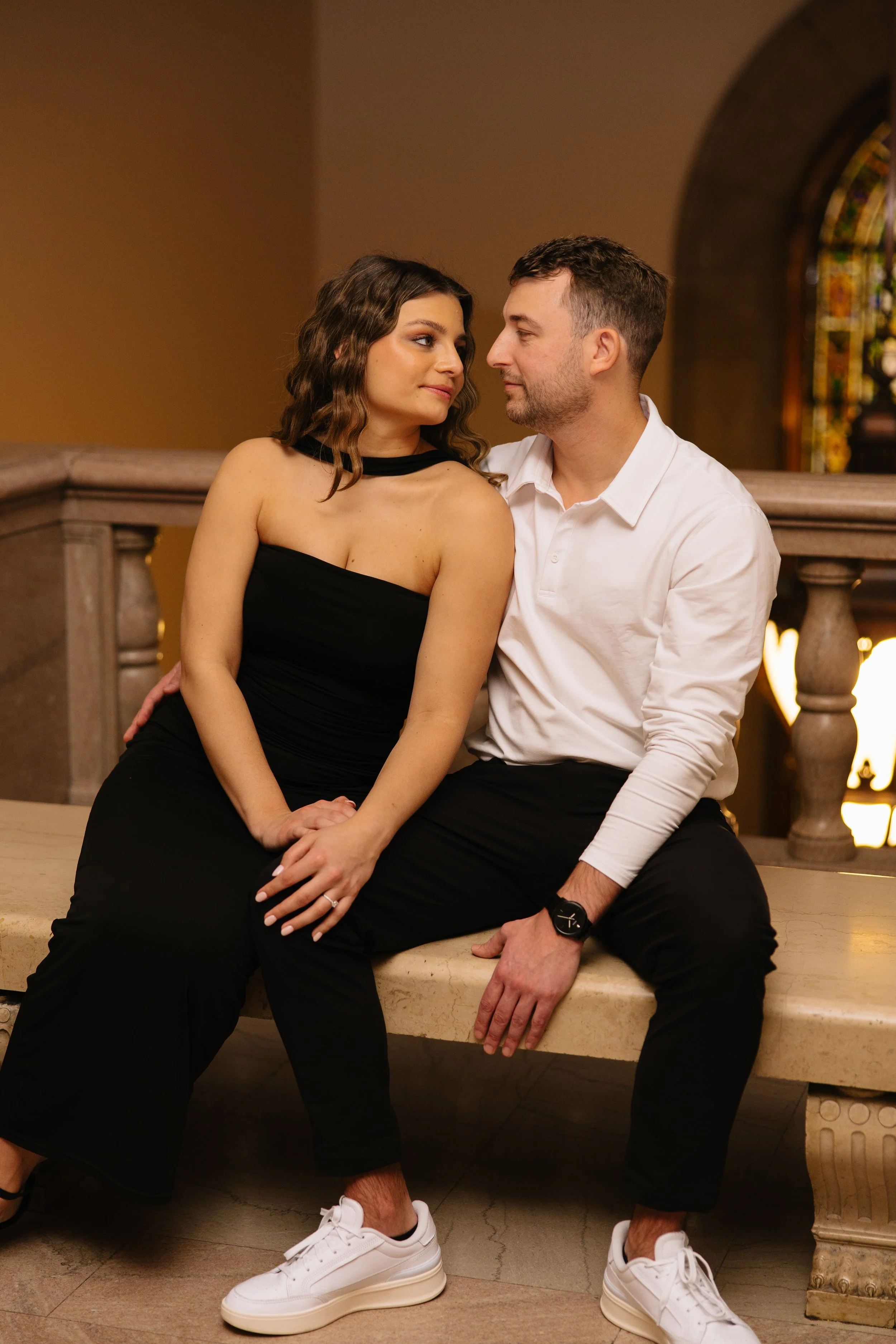 A couple sitting on a ledge inside a church, looking into each other's eyes. The woman wears a black off-the-shoulder dress, and the man wears a white shirt and black pants. They both wear white sneakers.