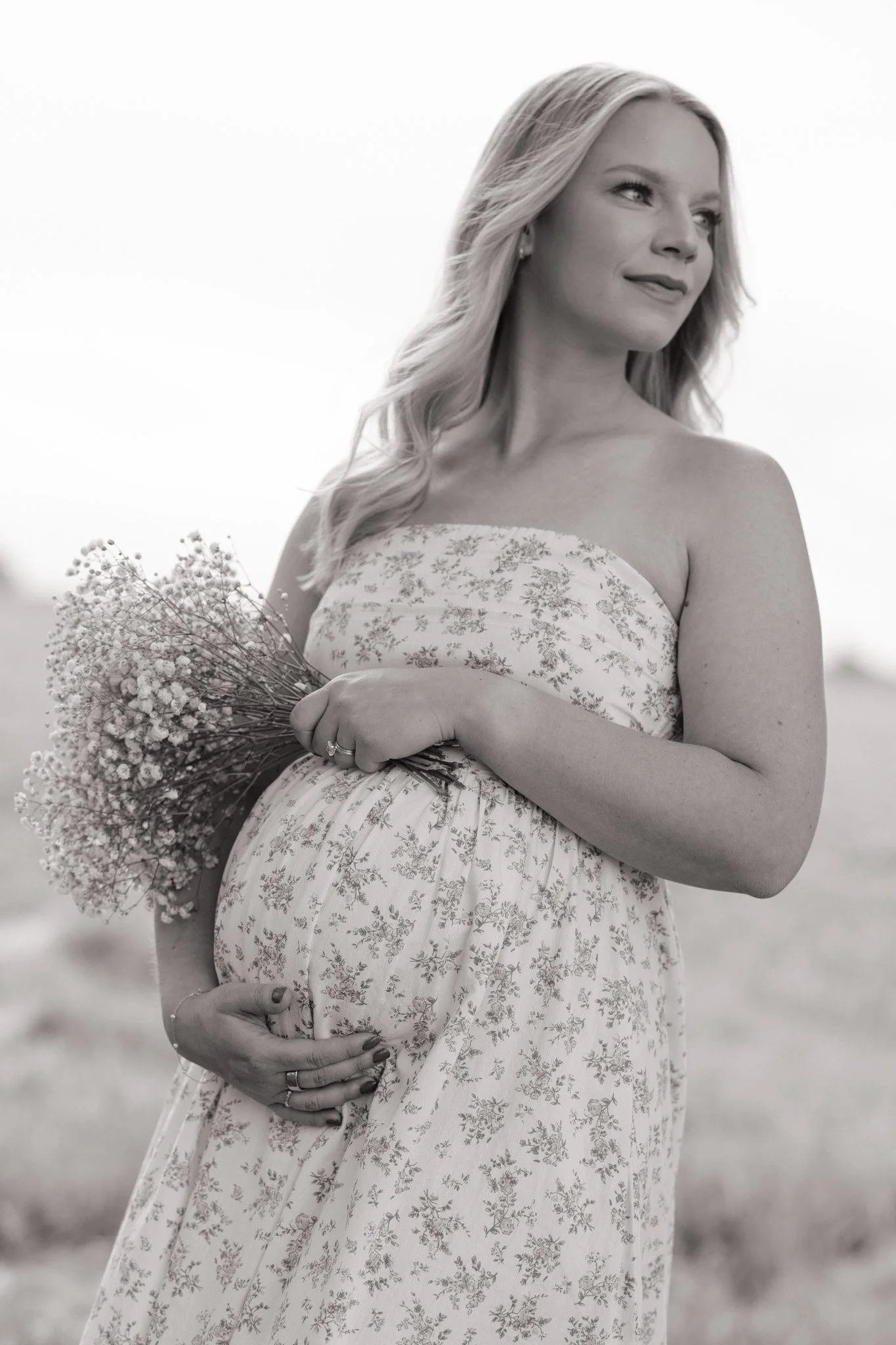 A pregnant woman with long wavy hair, wearing a strapless dress with a floral pattern, holding a bouquet of small flowers and her belly, standing outdoors in a natural setting.