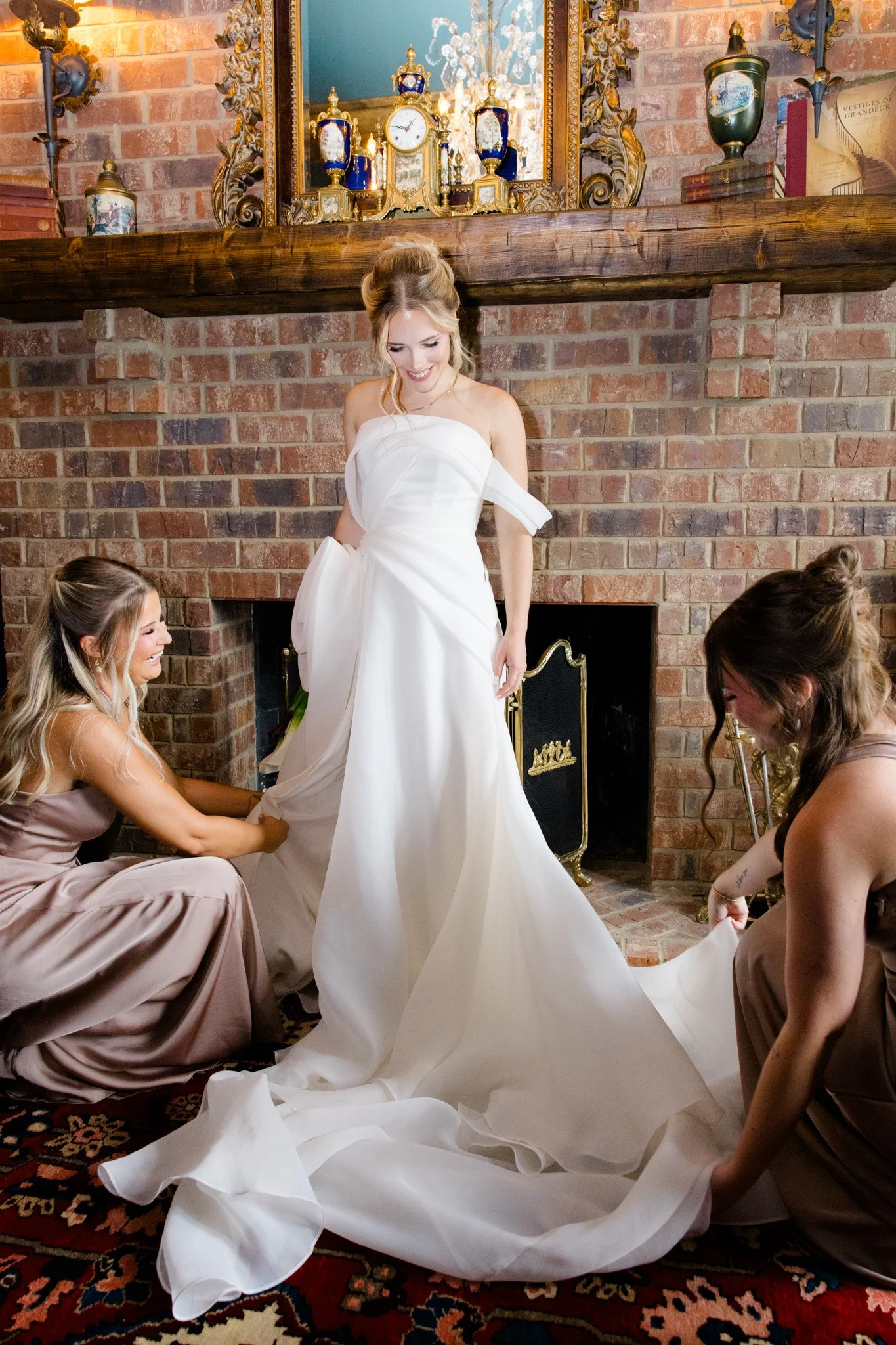 A bride in a white wedding dress stands in a living room with a brick fireplace and ornate mantle. She is surrounded by four women in taupe dresses assisting with her dress, which is being arranged or adjusted.