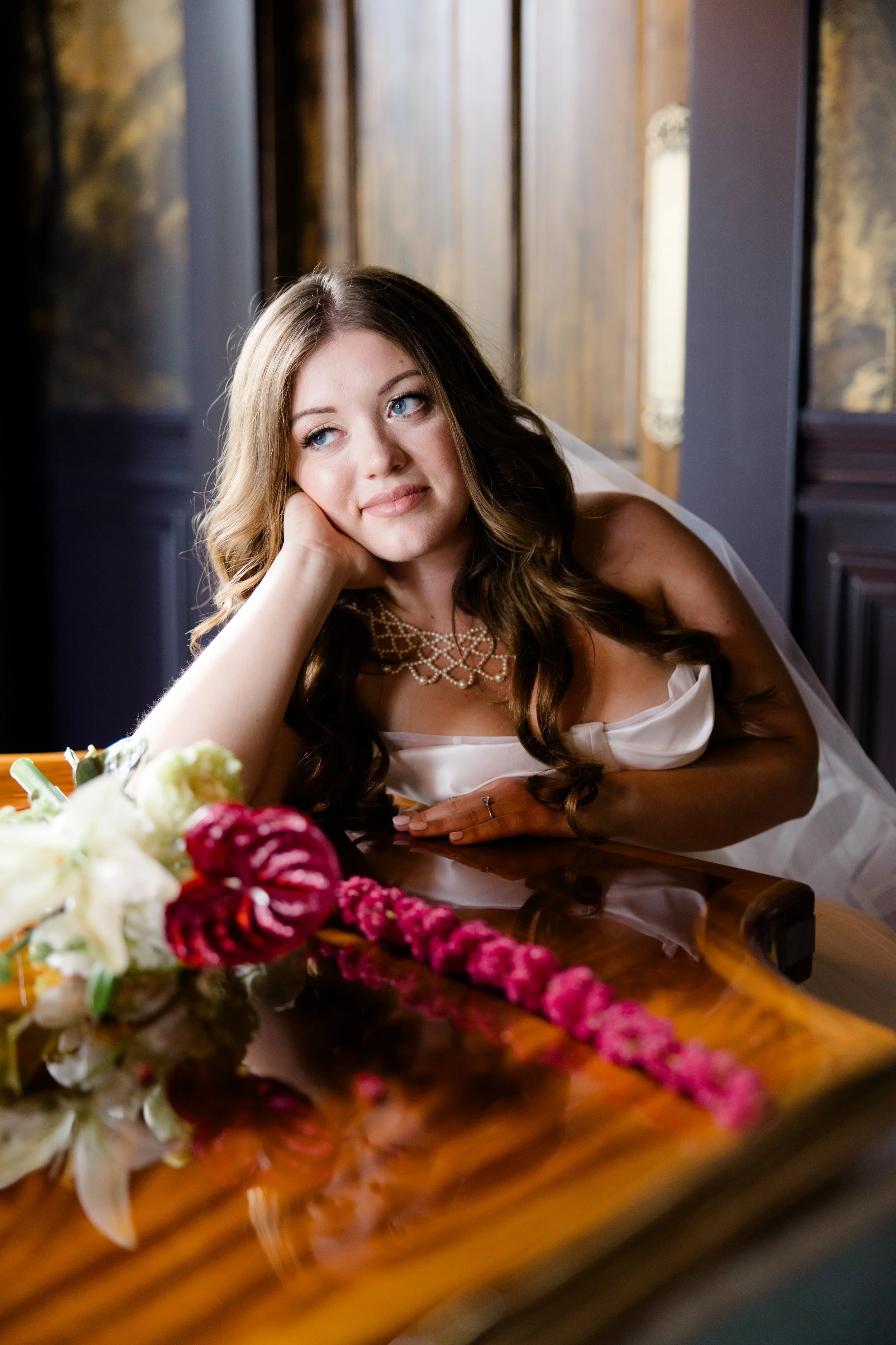 A woman with blue eyes and long brown hair, wearing a white dress and a pearl necklace, rests her head on her hand at a table with a floral centerpiece featuring white and pink flowers.