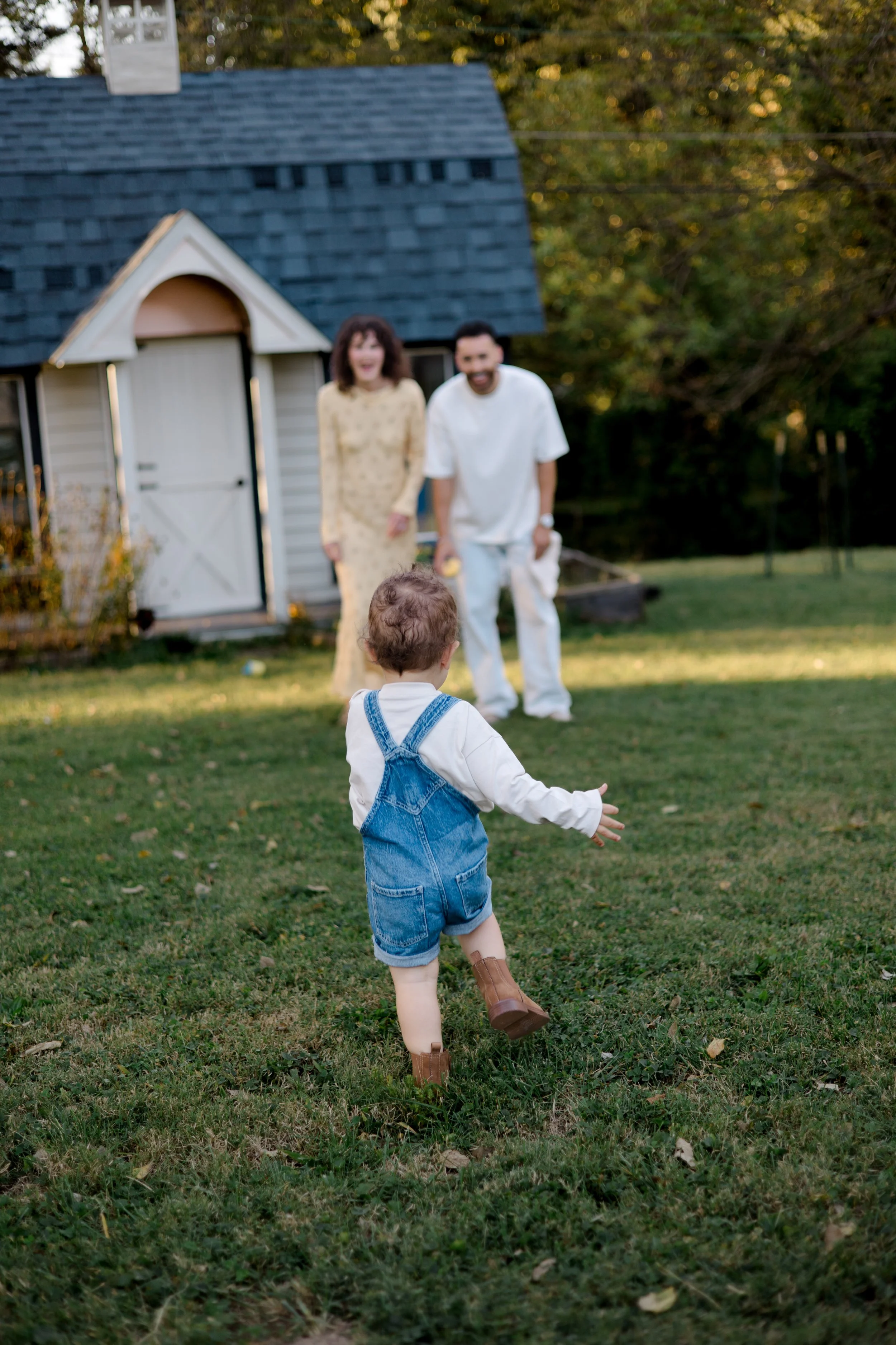 A young child wearing denim overalls and cowboy boots is running on a grassy lawn toward two adults in the background, who are holding hands and smiling in front of a small house with a blue roof.