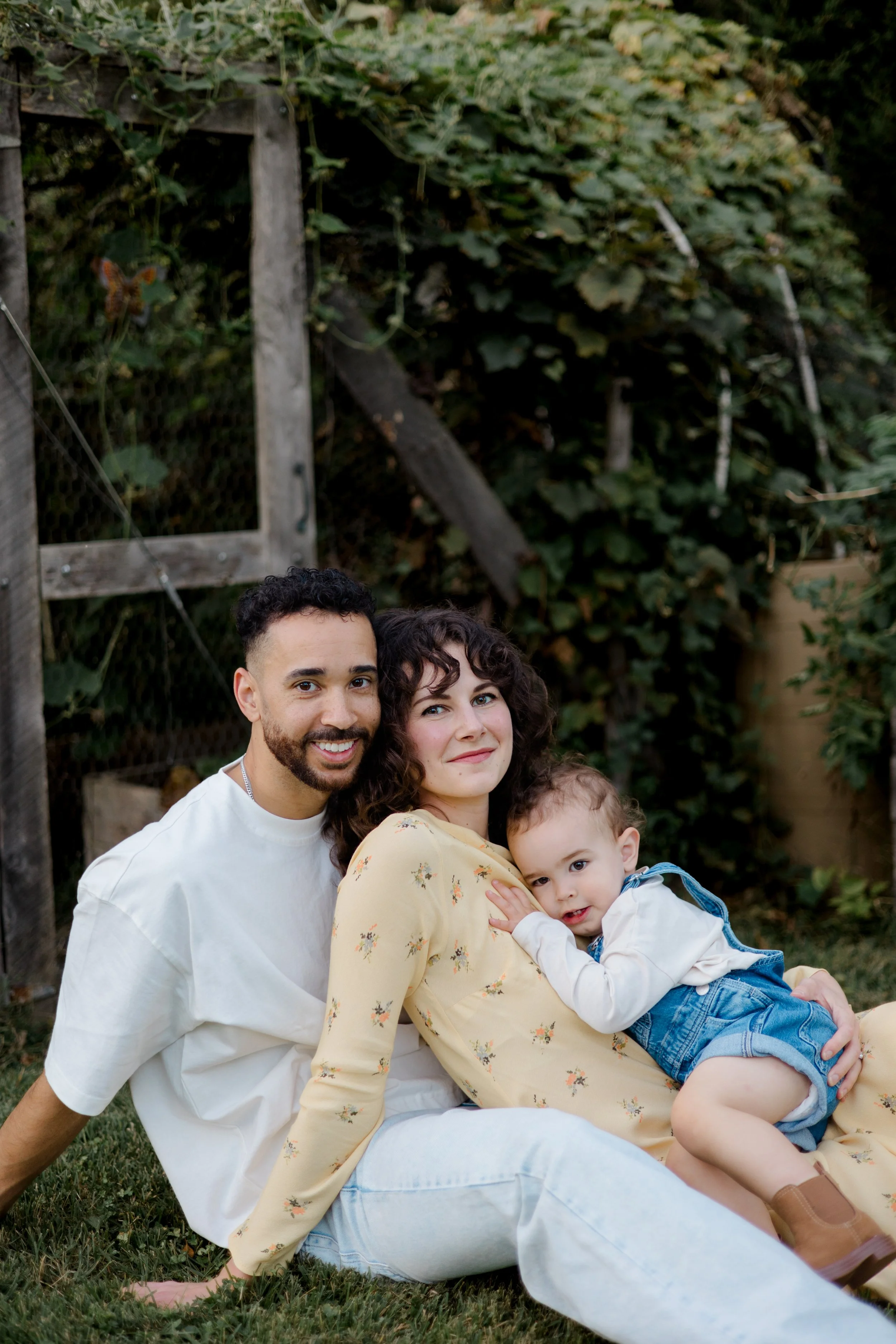 A happy multicultural family of three sitting outdoors on grass, with two adults and a young child, smiling at the camera.