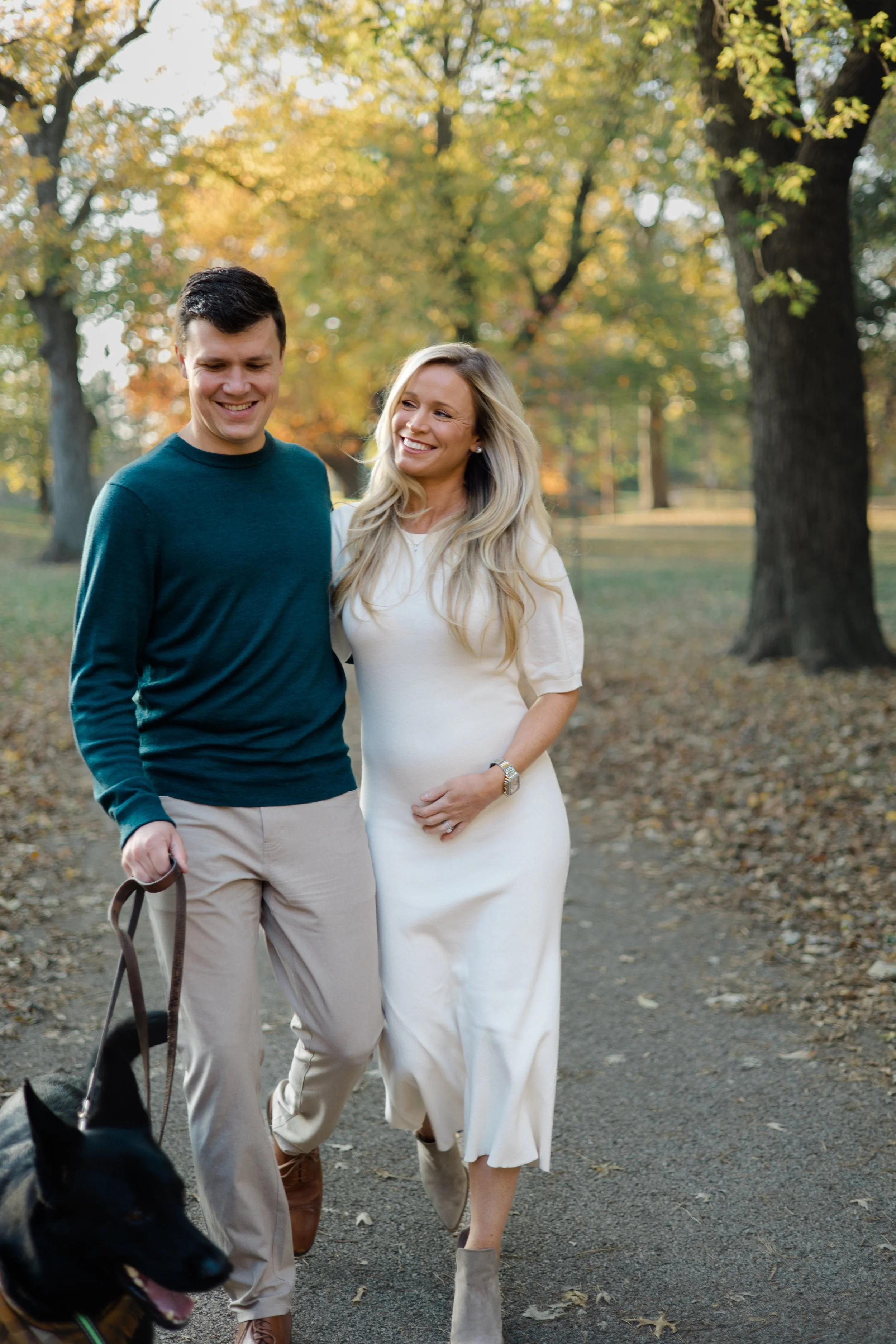 A couple walking outdoors in a park during autumn with trees and fallen leaves around. The man is wearing a dark green long-sleeve shirt and beige pants, holding a leash with a black dog. The woman is wearing a white dress and light gray ankle boots,