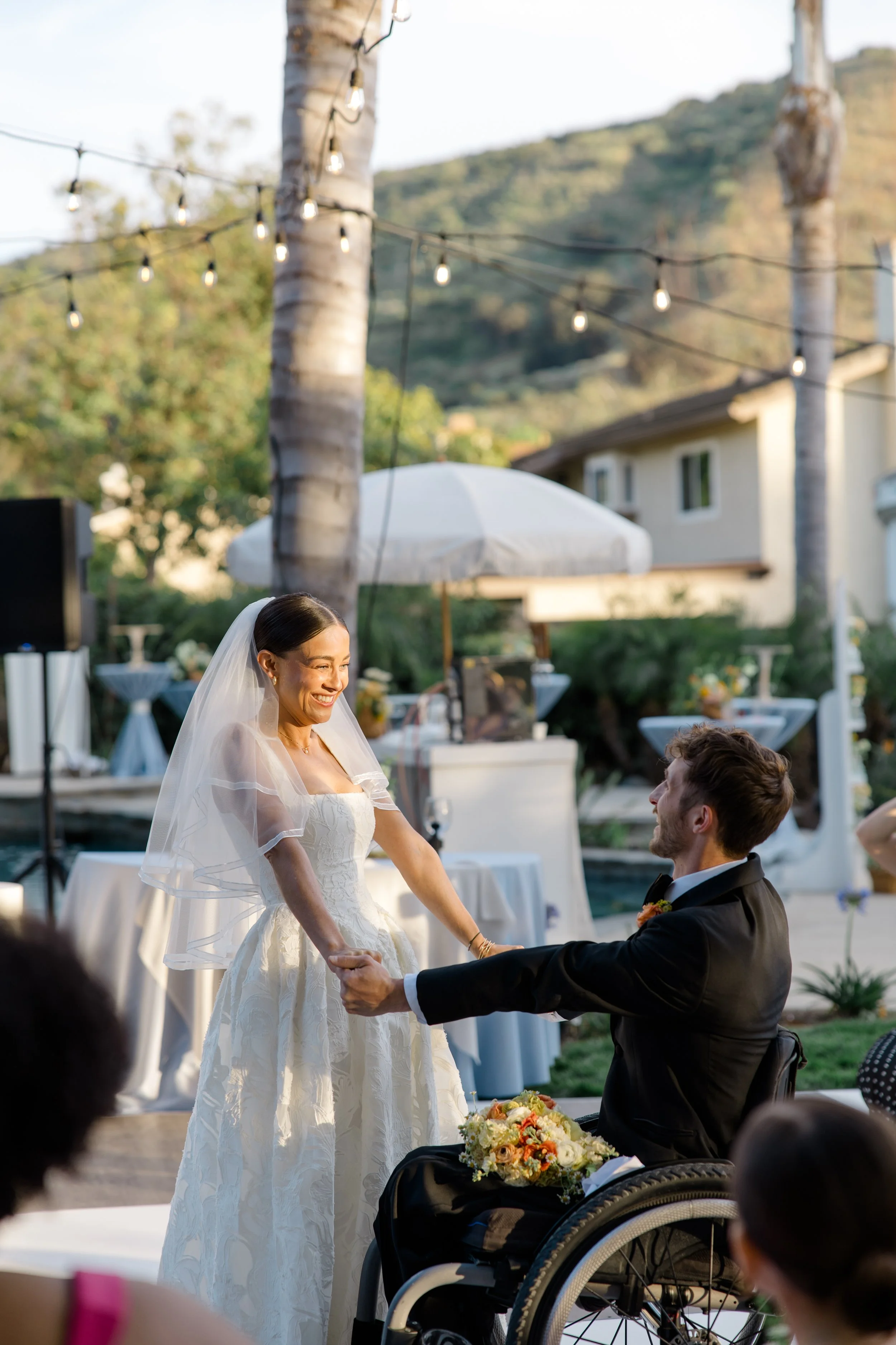 A bride and groom exchange vows outdoors at a wedding ceremony, with the bride wearing a white wedding dress and veil, and the groom in a black suit, seated in a wheelchair. They are surrounded by guests, with tables and string lights overhead, and a