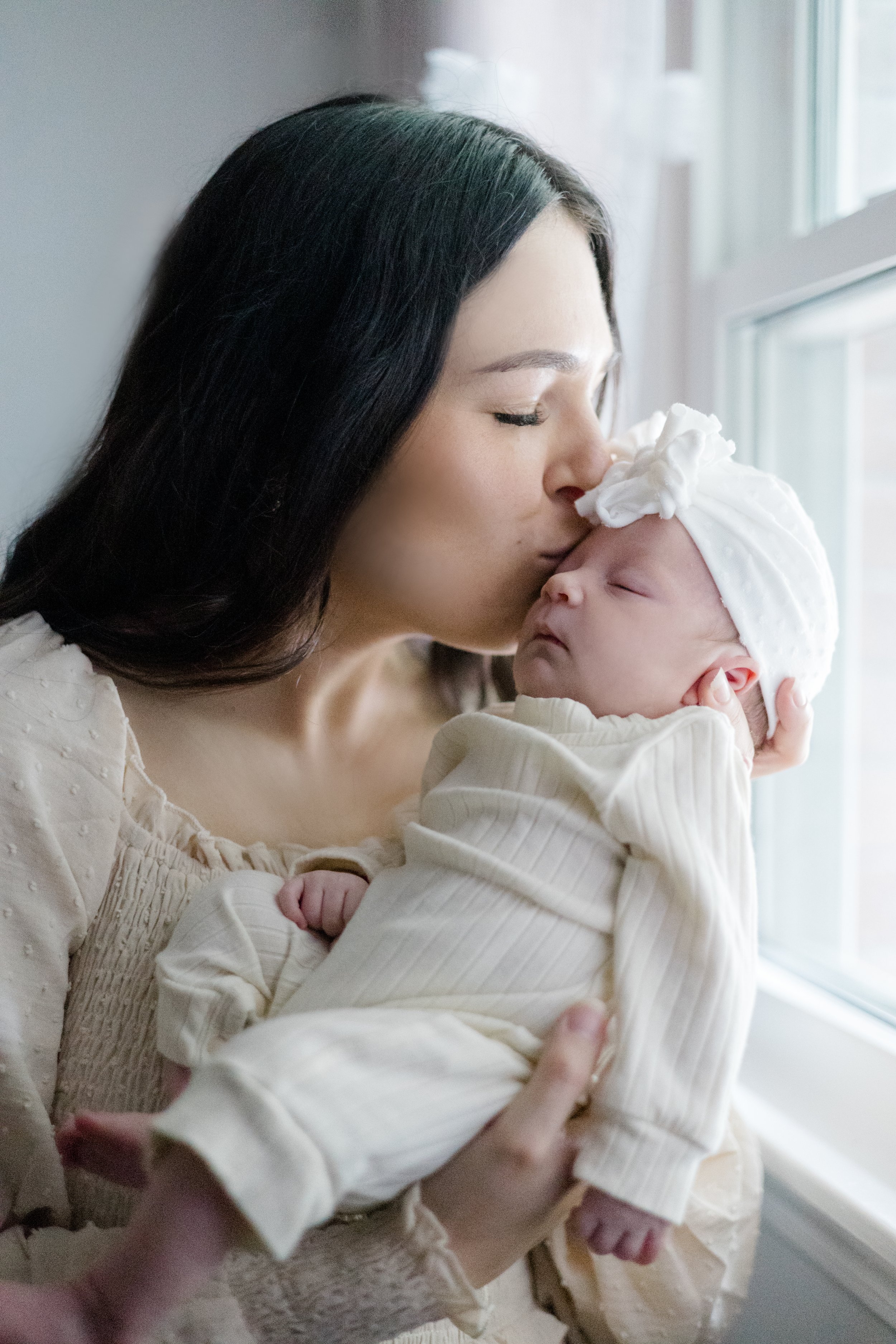 A woman with dark hair and light skin gently holding a sleeping baby dressed in cream-colored clothing and a matching hat, near a window with natural light.