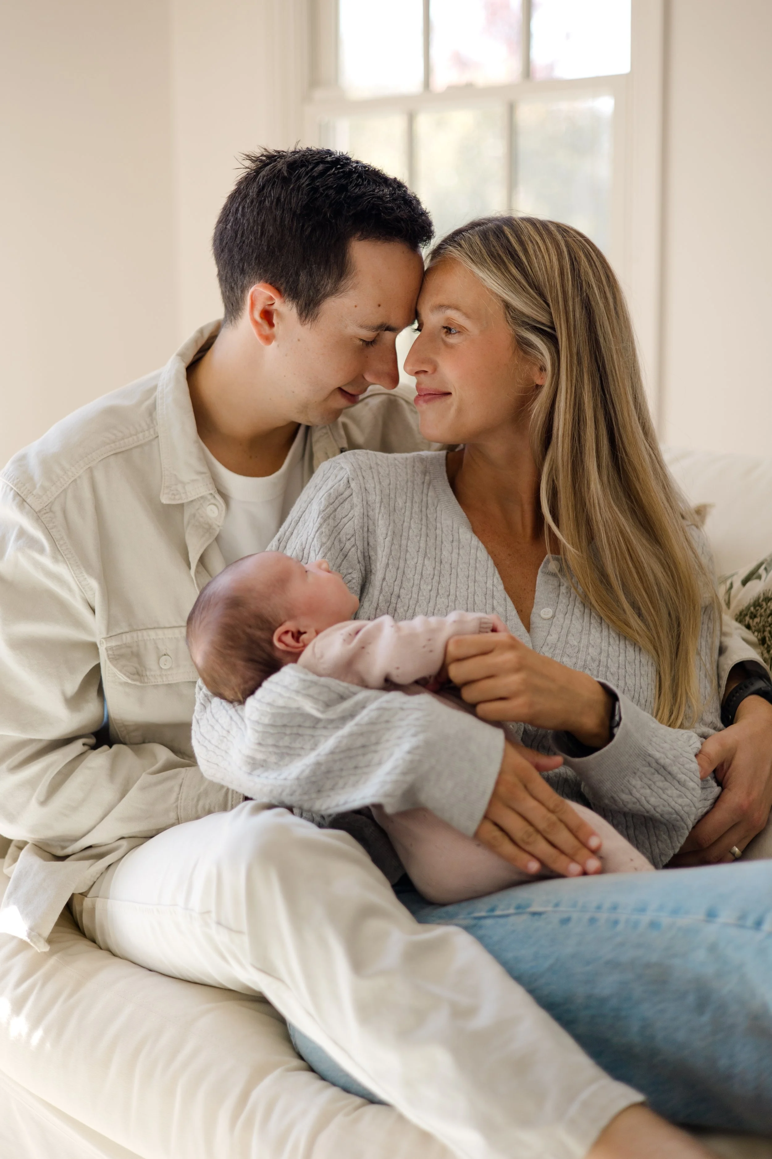 A family with a newborn baby in a cozy living room. The mother and father are sitting close, with their foreheads touching, showing love and affection. The mother is holding the baby, who is resting peacefully in her arms. Natural light is coming thr