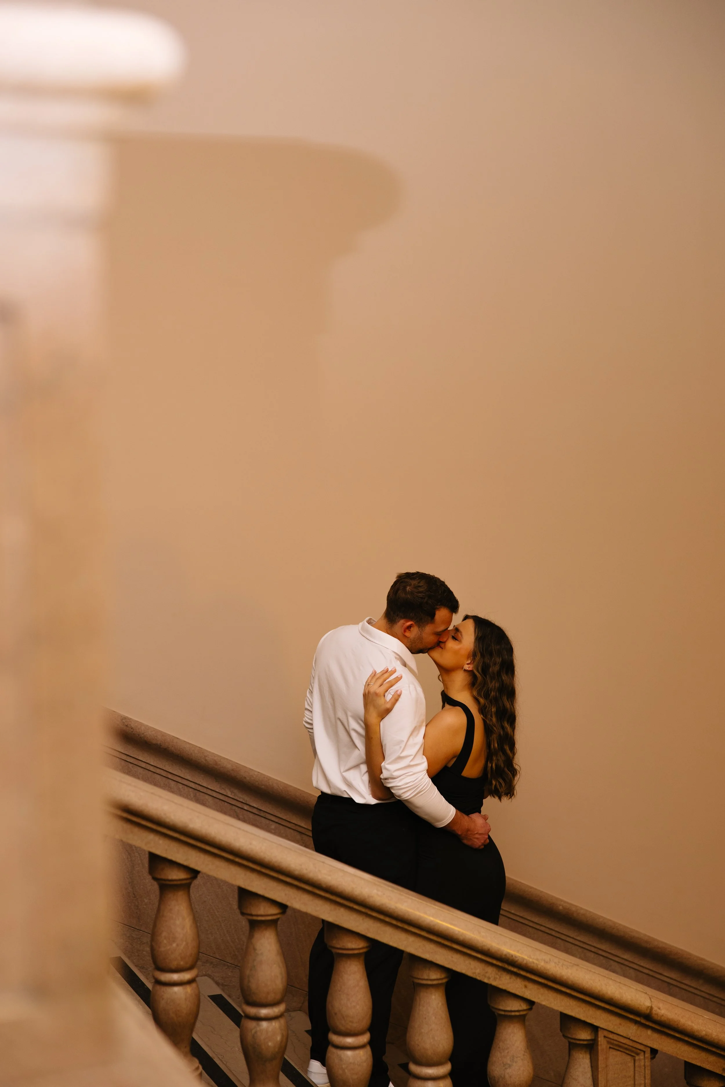 A couple in formal attire sharing a kiss on a staircase in an elegant interior setting.