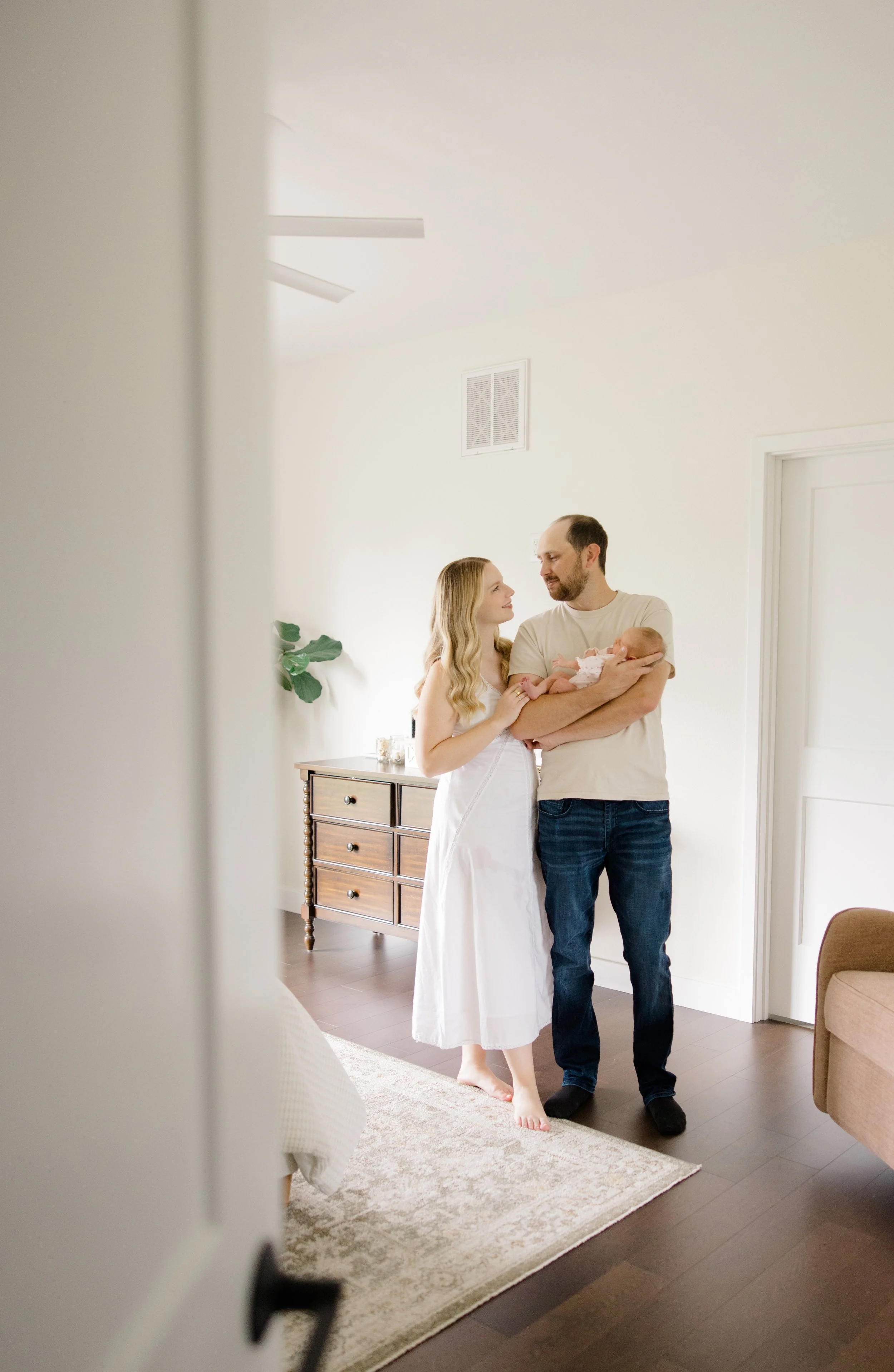 A couple standing in a bedroom holding a baby, with a white wall and wooden dresser in the background.