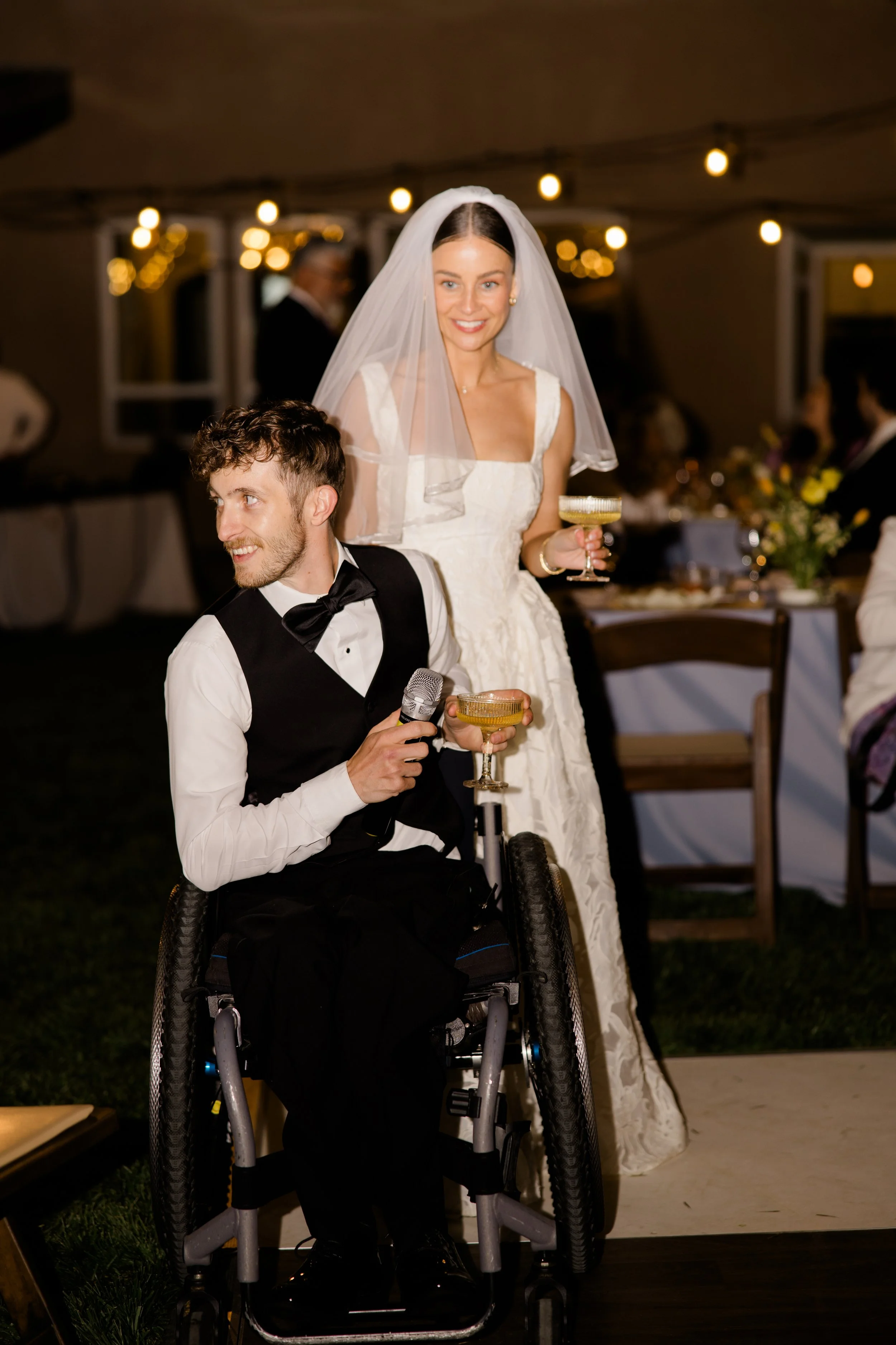 A bride in a wedding gown and veil holding a champagne glass stands next to a man in a wheelchair wearing a tuxedo at a wedding reception.