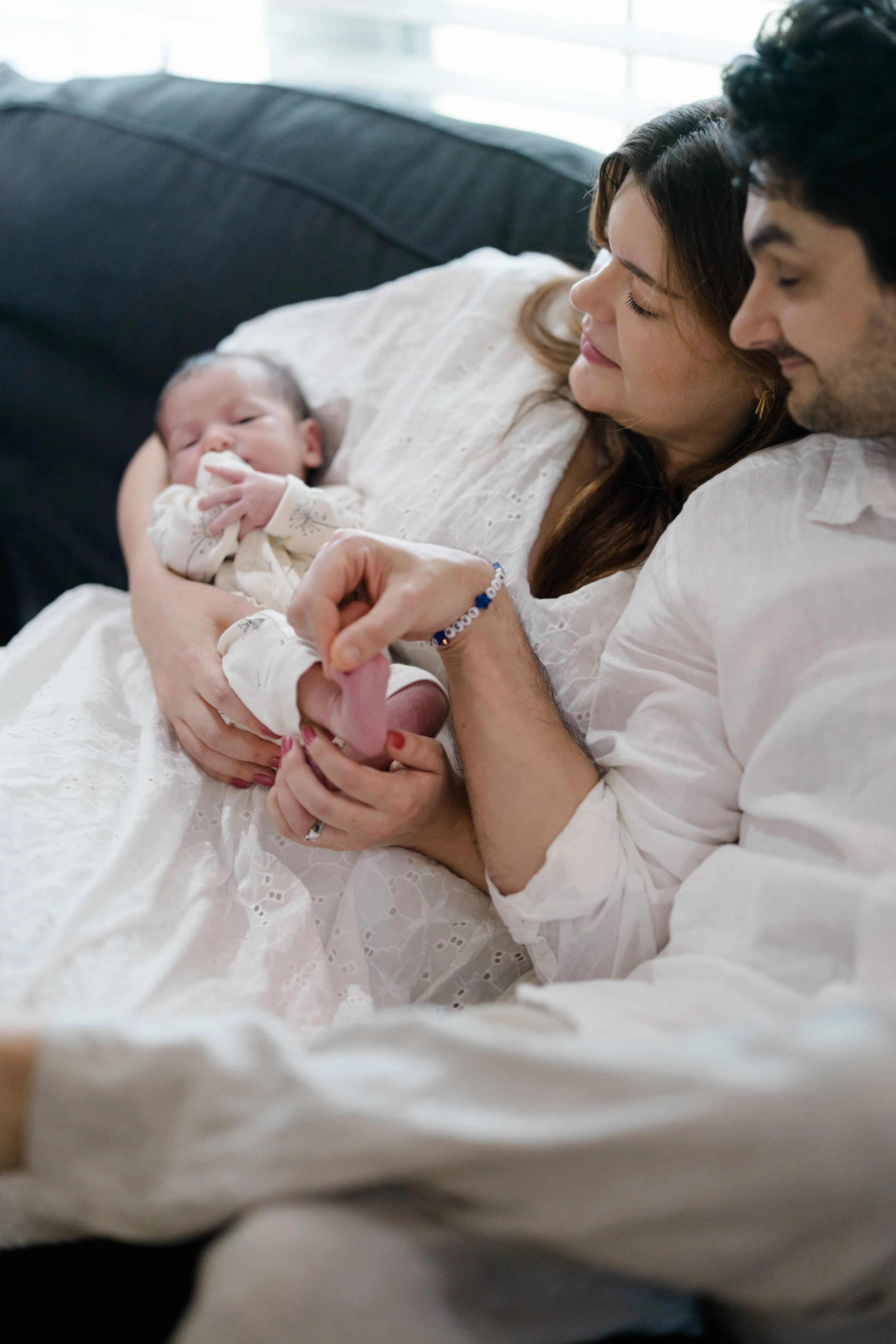 A woman, man, and baby lying on a couch. The woman is holding the baby on her chest, and the man is holding the baby's hand while smiling at the baby.