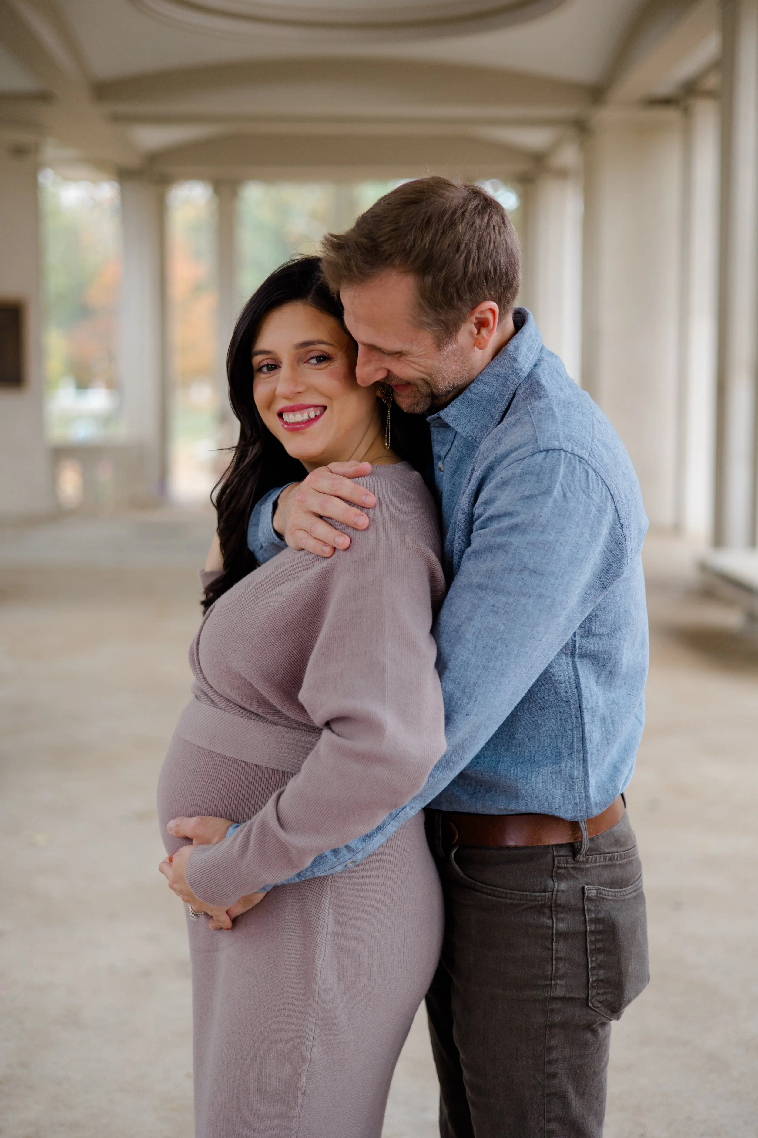 A pregnant woman in a beige dress smiling while hugging a man in a blue shirt, both standing in an outdoor covered area with a blurred background.