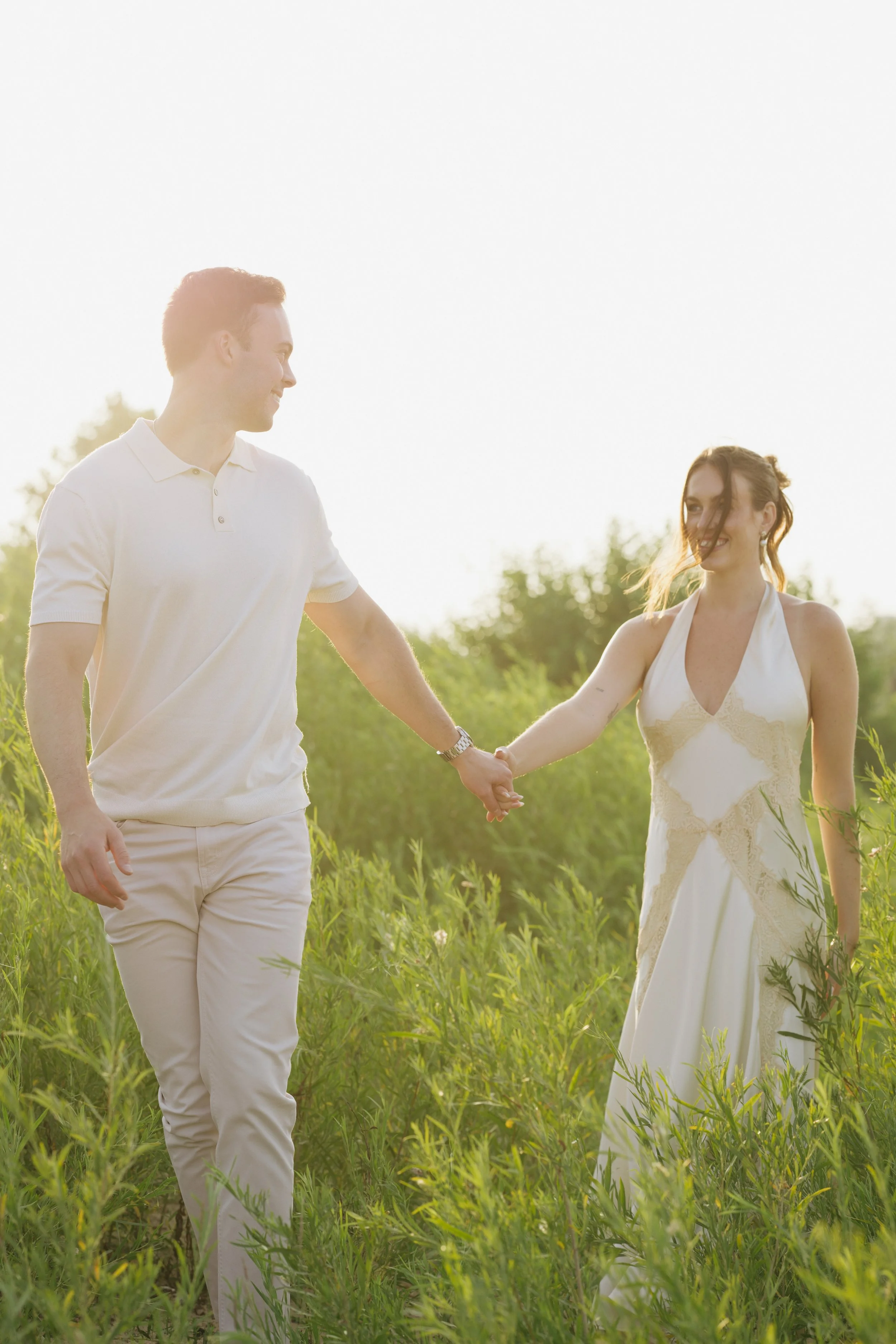A man and woman holding hands and walking through a field of green plants during sunset.