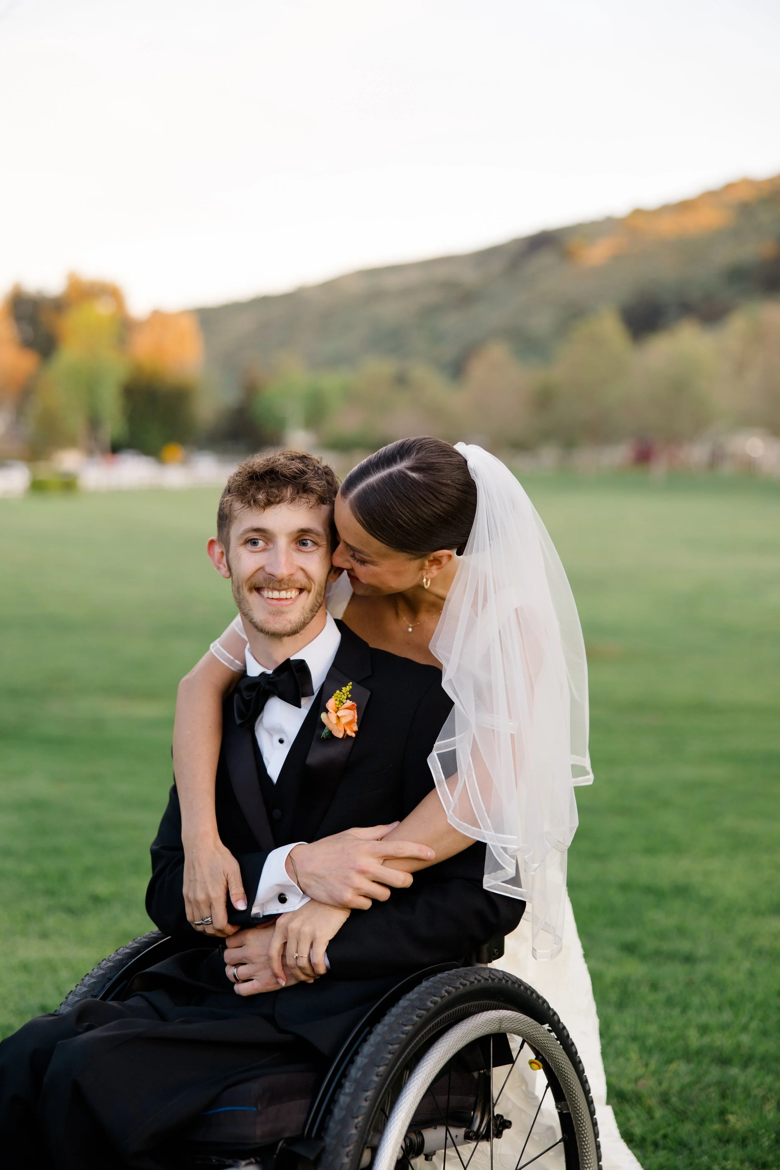 A newlywed couple; the groom, a man with curly brown hair and beard in a wheelchair, wearing a black tuxedo, and the bride, a woman with dark hair styled in a slicked-back ponytail, smiling, holding a bouquet of flowers, leaning in to kiss him on the