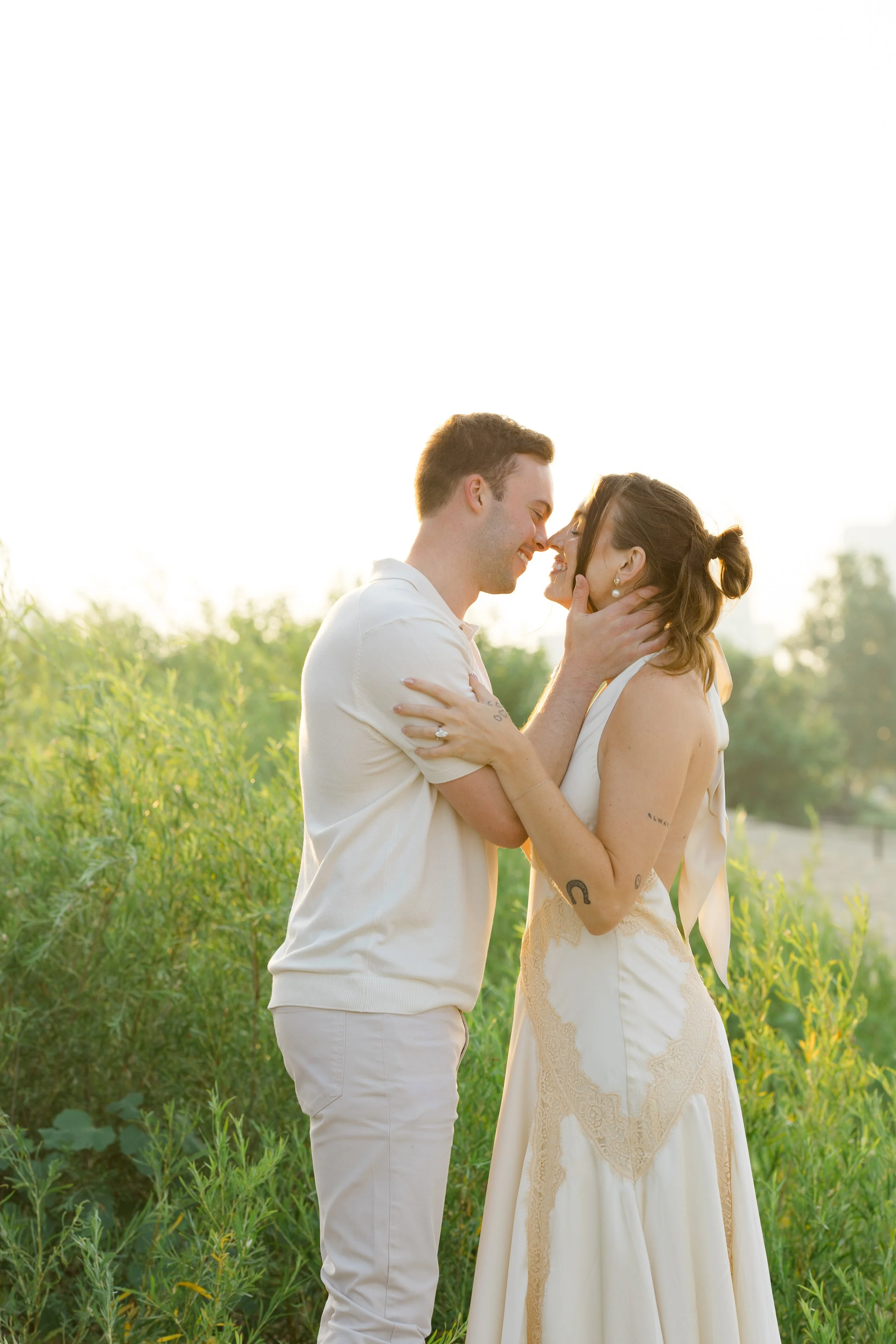 A couple standing close together outdoors, smiling and touching noses, surrounded by greenery during sunset.