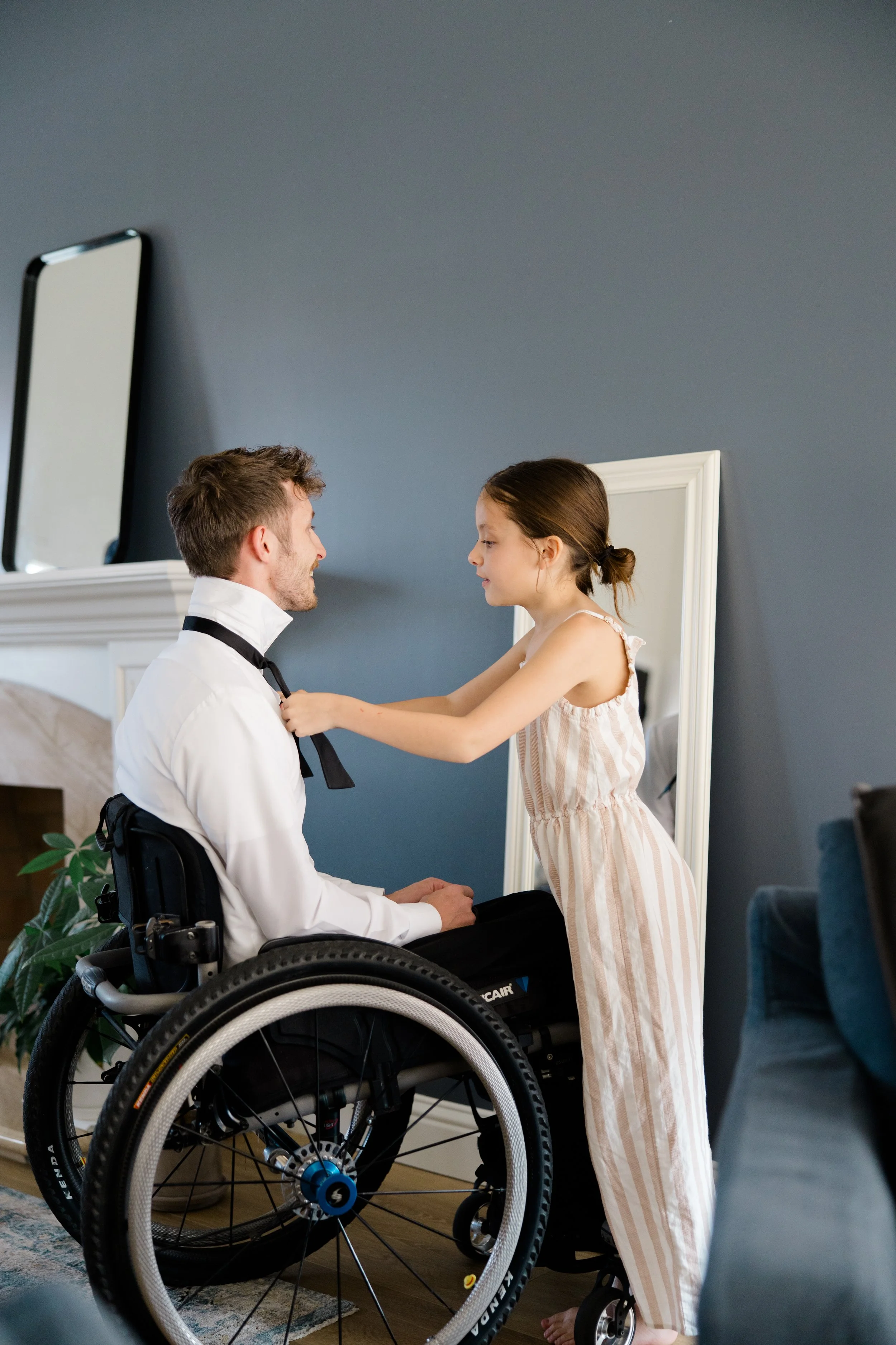 A young girl in striped pajamas standing in front of a man in a wheelchair, adjusting his bow tie as they look at each other in a room with a mirror and a blue wall.