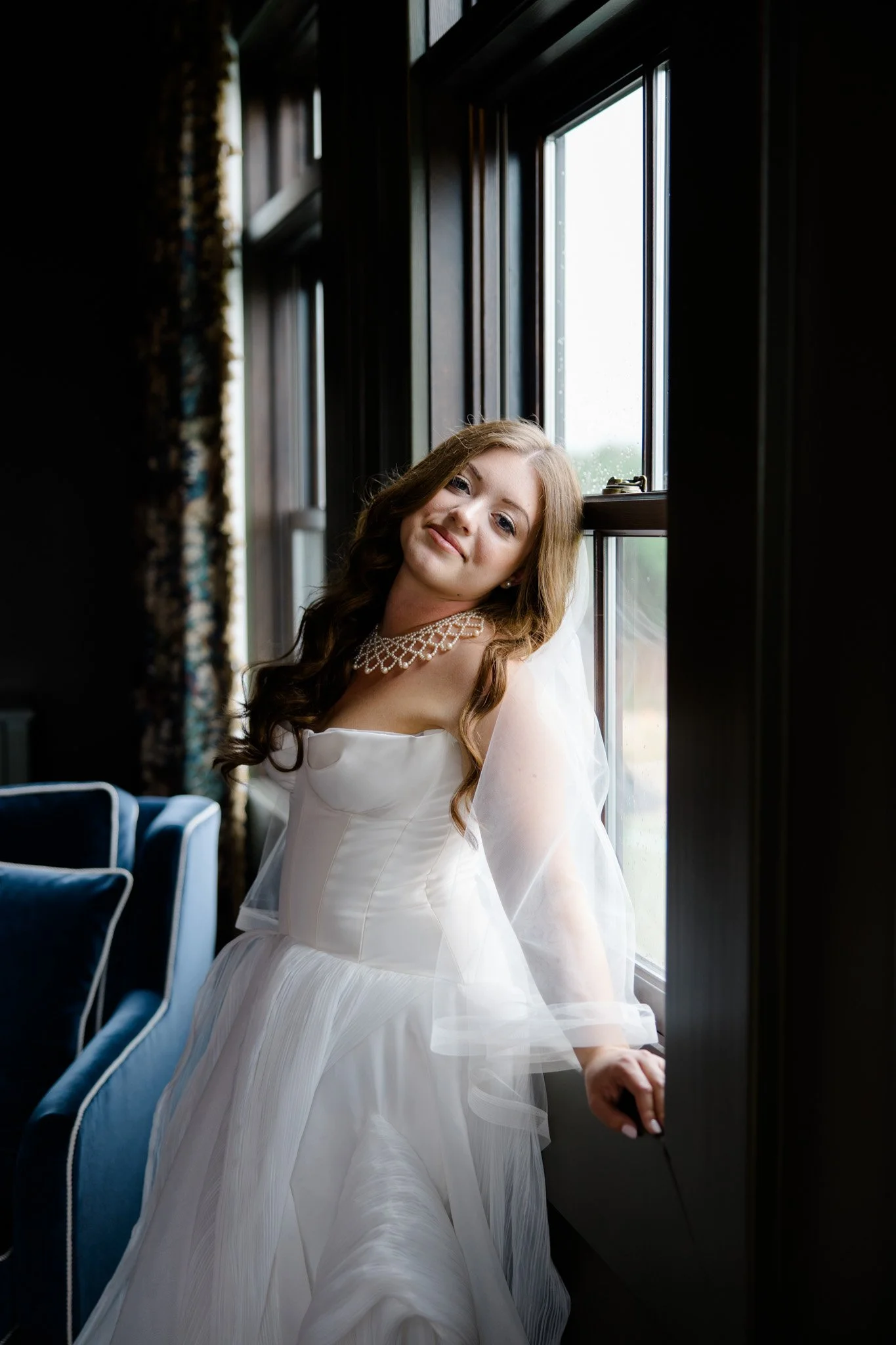 A young woman in a white wedding dress sitting by a window, with her head resting on the window frame, in a dimly lit room.
