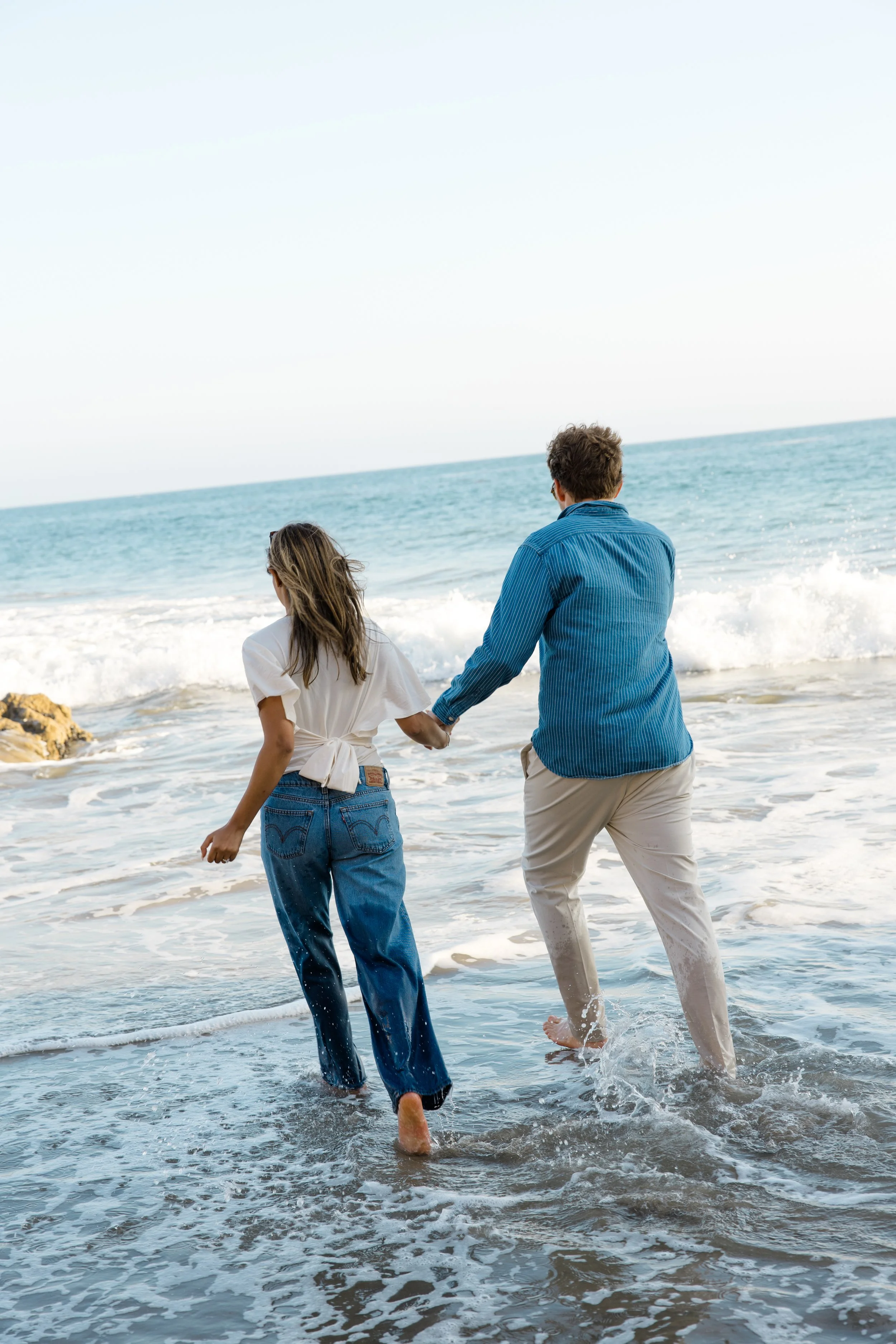 A man and woman holding hands and walking in the shallow surf at the beach, with waves crashing around their feet and the ocean extending to the horizon.